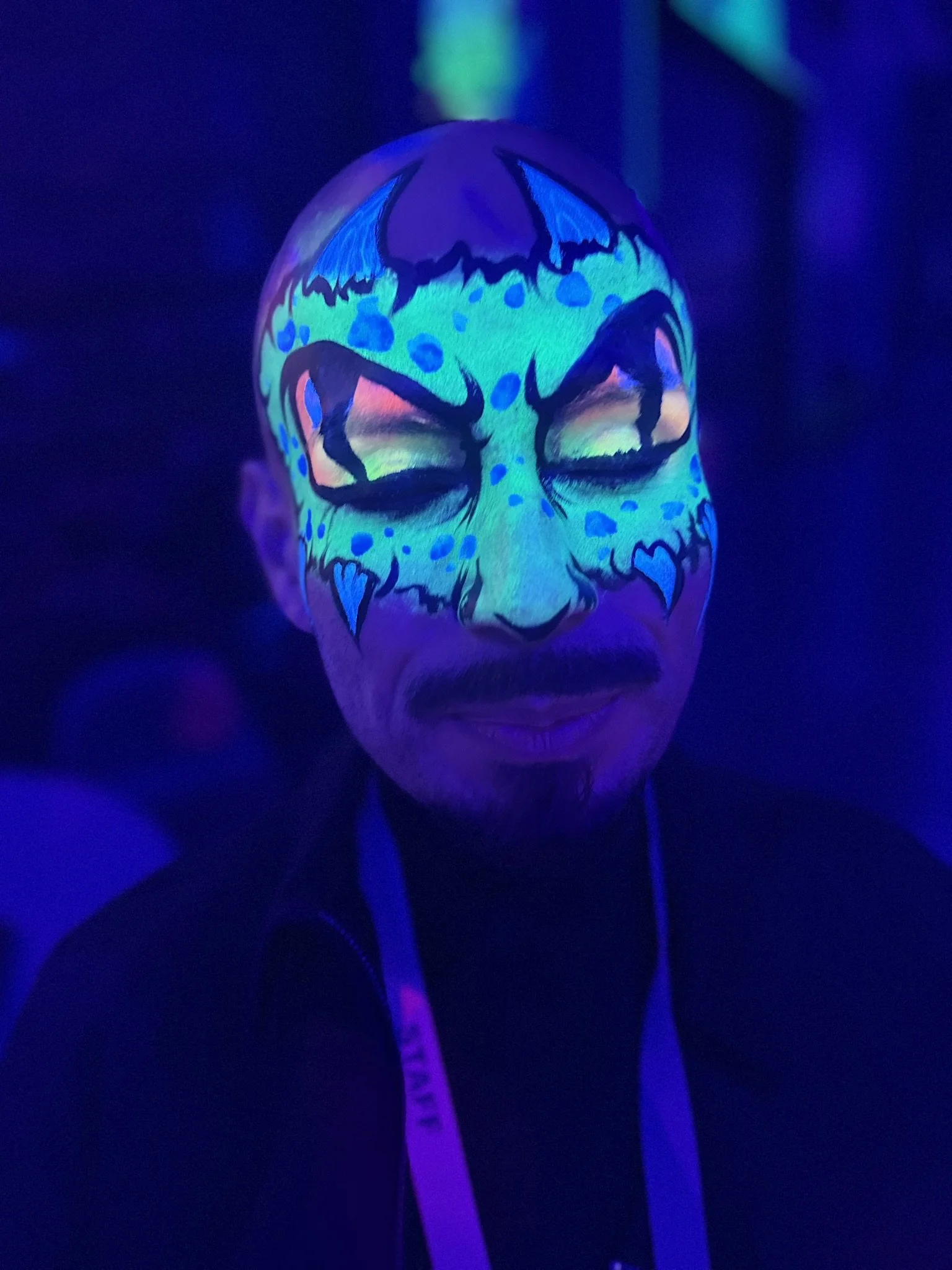 A person with glowing face paint under UV light, designed to look like a monster with exaggerated features and bright colors, wearing a lanyard.