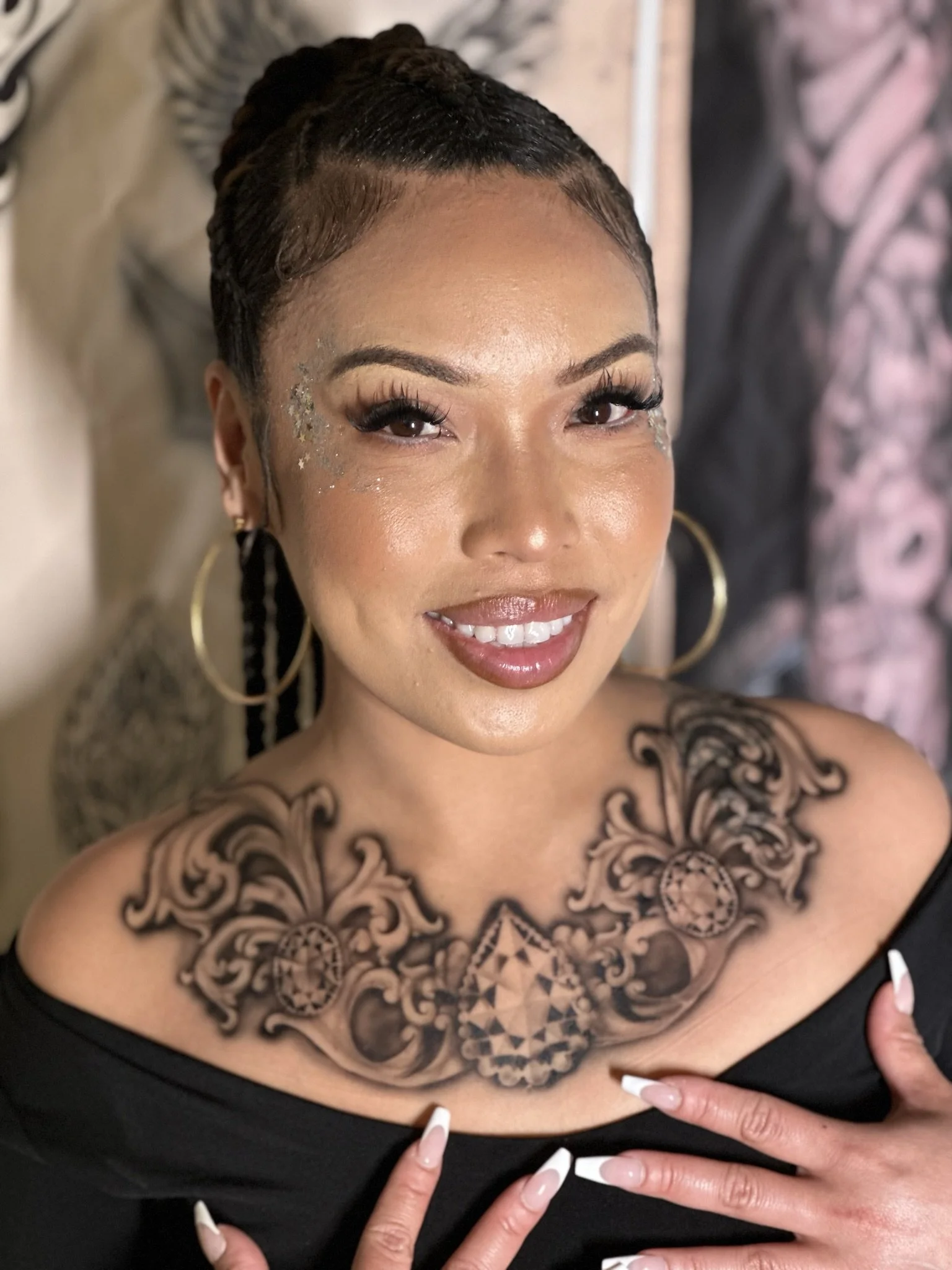 Woman with braided hairstyle and decorative chest tattoo, wearing hoop earrings and glitter makeup.