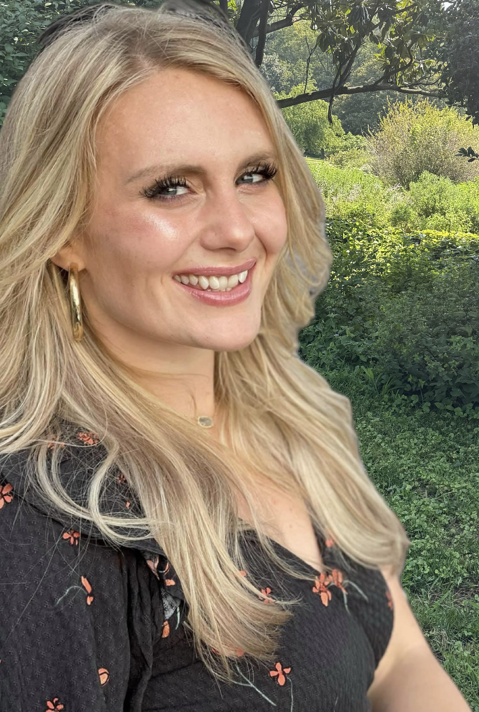 A woman with long blonde hair, wearing earrings and a black top with red floral patterns, smiling outdoors in a lush green garden with trees and bushes in the background.