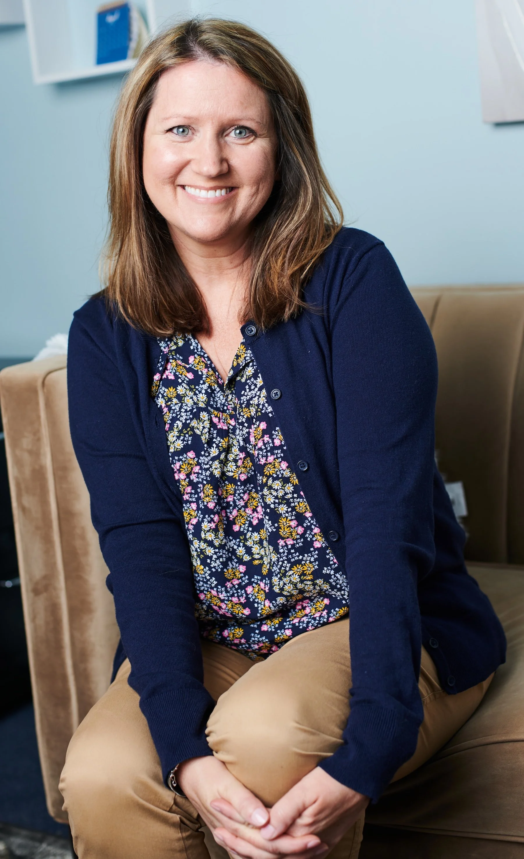 A woman with shoulder-length brown hair and blue eyes sitting on a beige sofa, smiling, wearing a navy cardigan over a floral blouse and tan pants.
