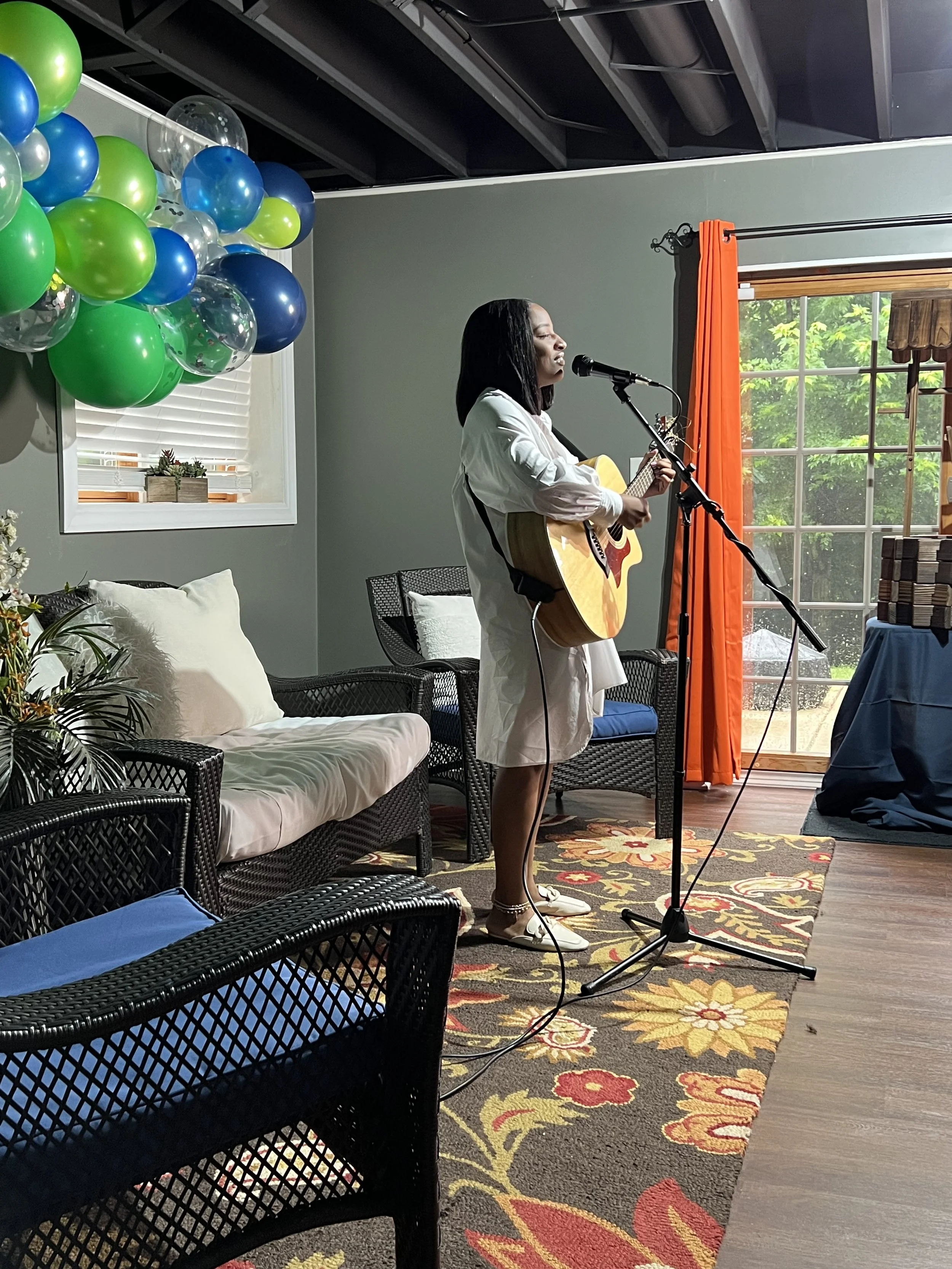 A woman in a white dress is performing a song with a guitar at a small indoor event, standing on a colorful floral carpet with a microphone. There are green, blue, silver, and transparent balloons on the left side and a window with orange curtains on