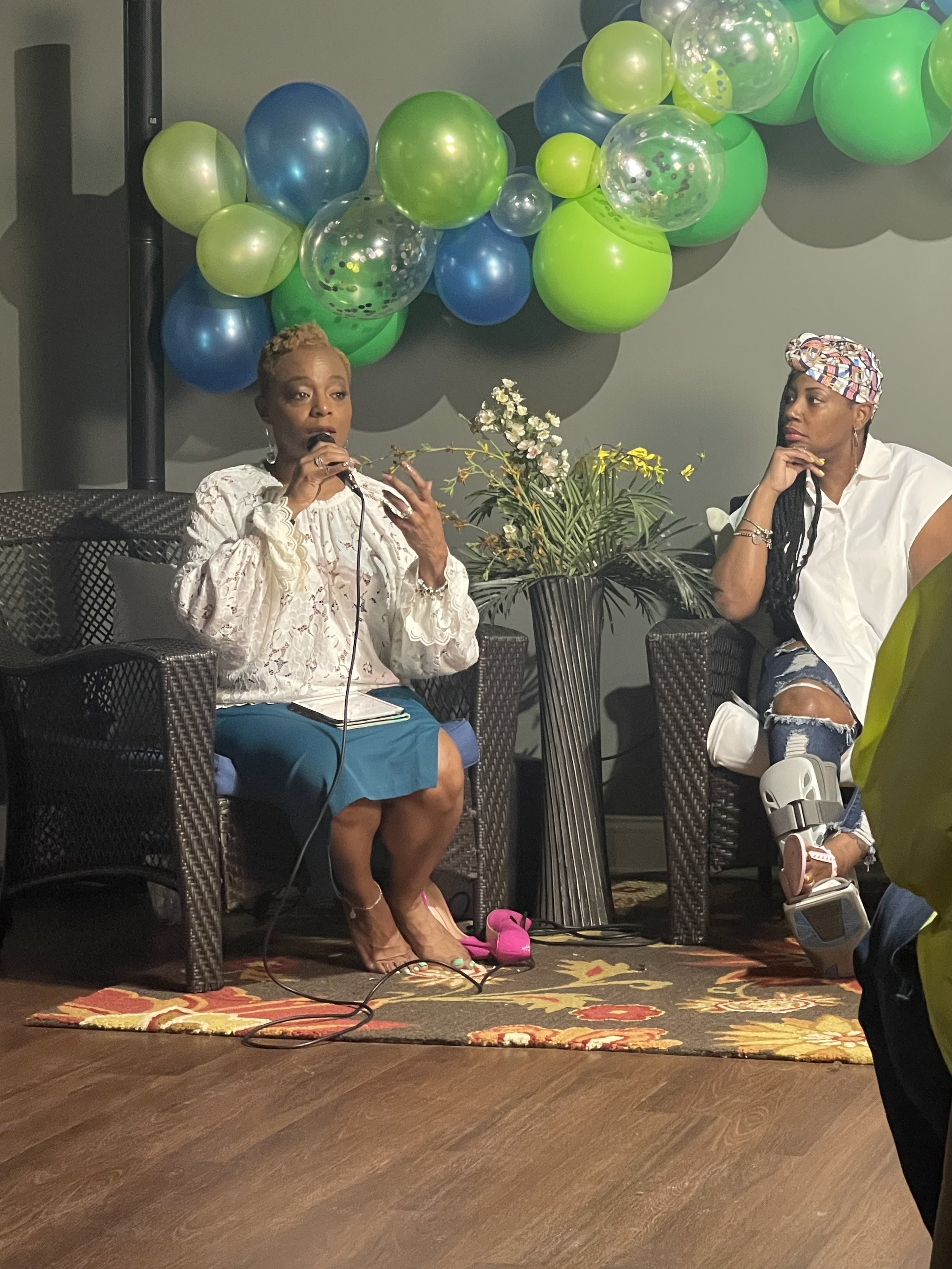 Two women seated in chairs, engaged in a conversation during an event, with colorful balloons and a floral arrangement in the background, and one woman speaking into a microphone.