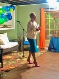 Woman standing and singing or speaking into a microphone in a decorated room with balloons, a couch, and a small stage area.