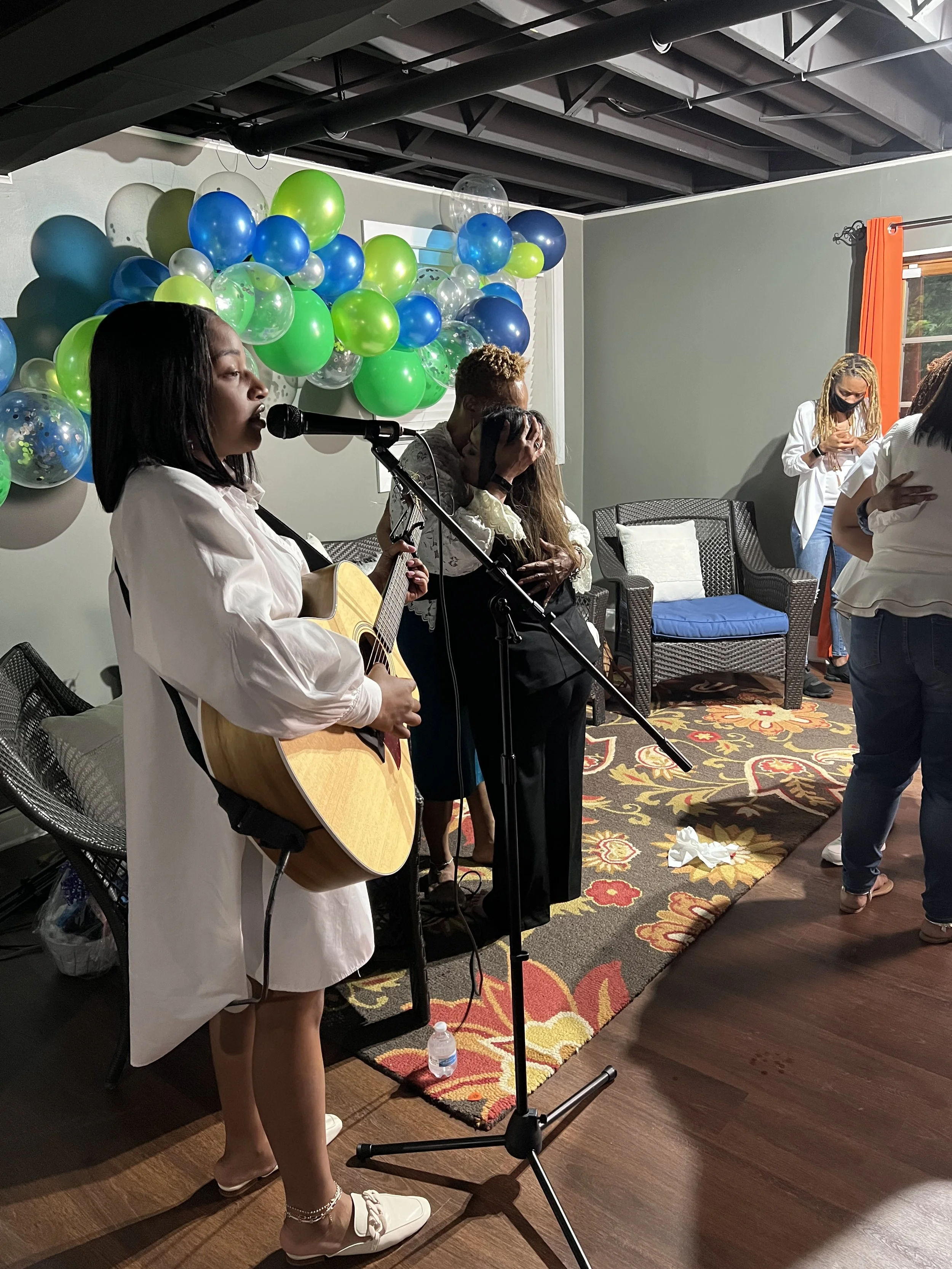 A woman singing and playing guitar during a celebration, with balloons in the background and other people in the room.