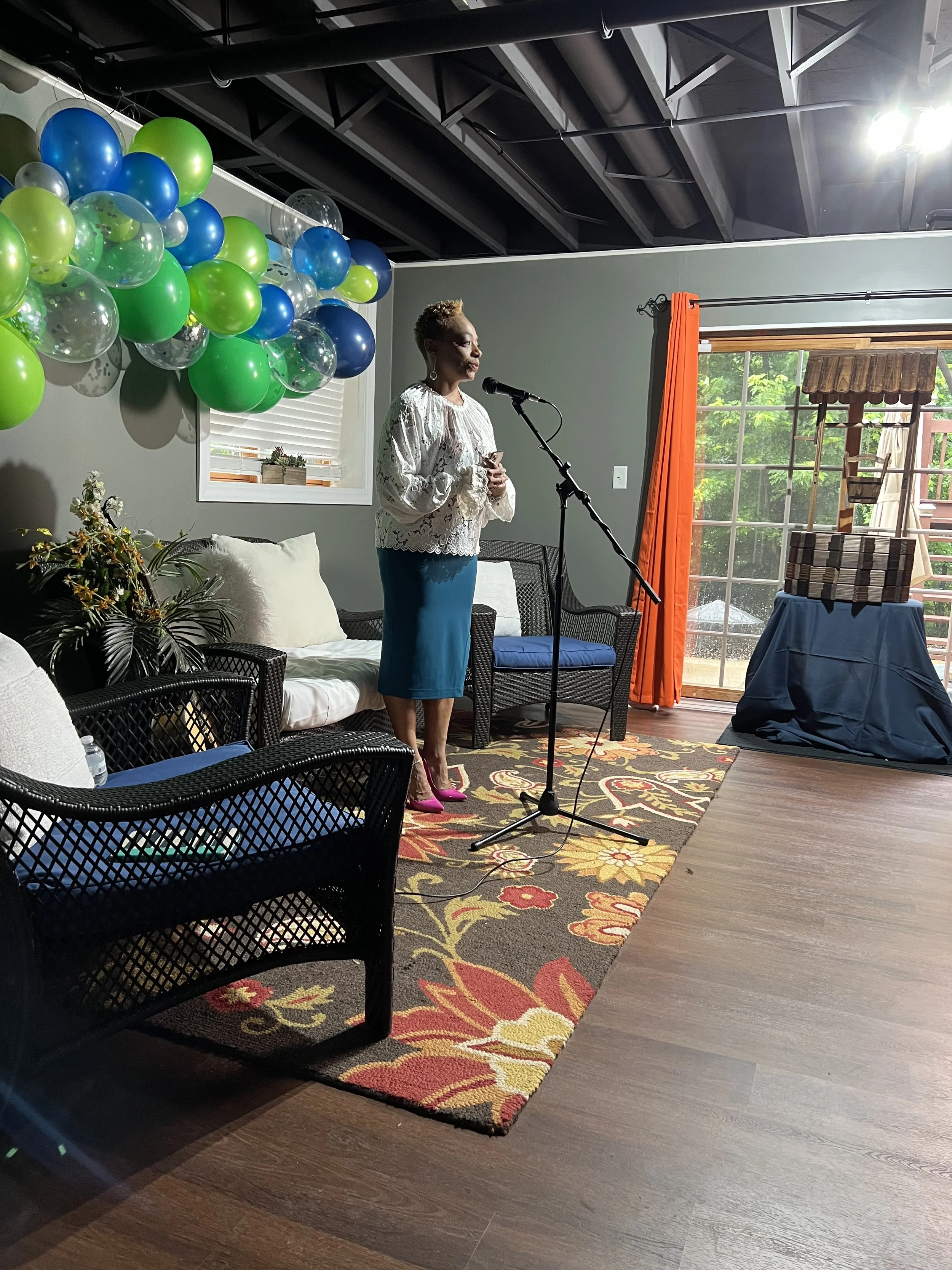 A woman speaking at a microphone in a decorated indoor space with balloons on the wall, a window with blinds, and a sliding glass door showing an outdoor patio.
