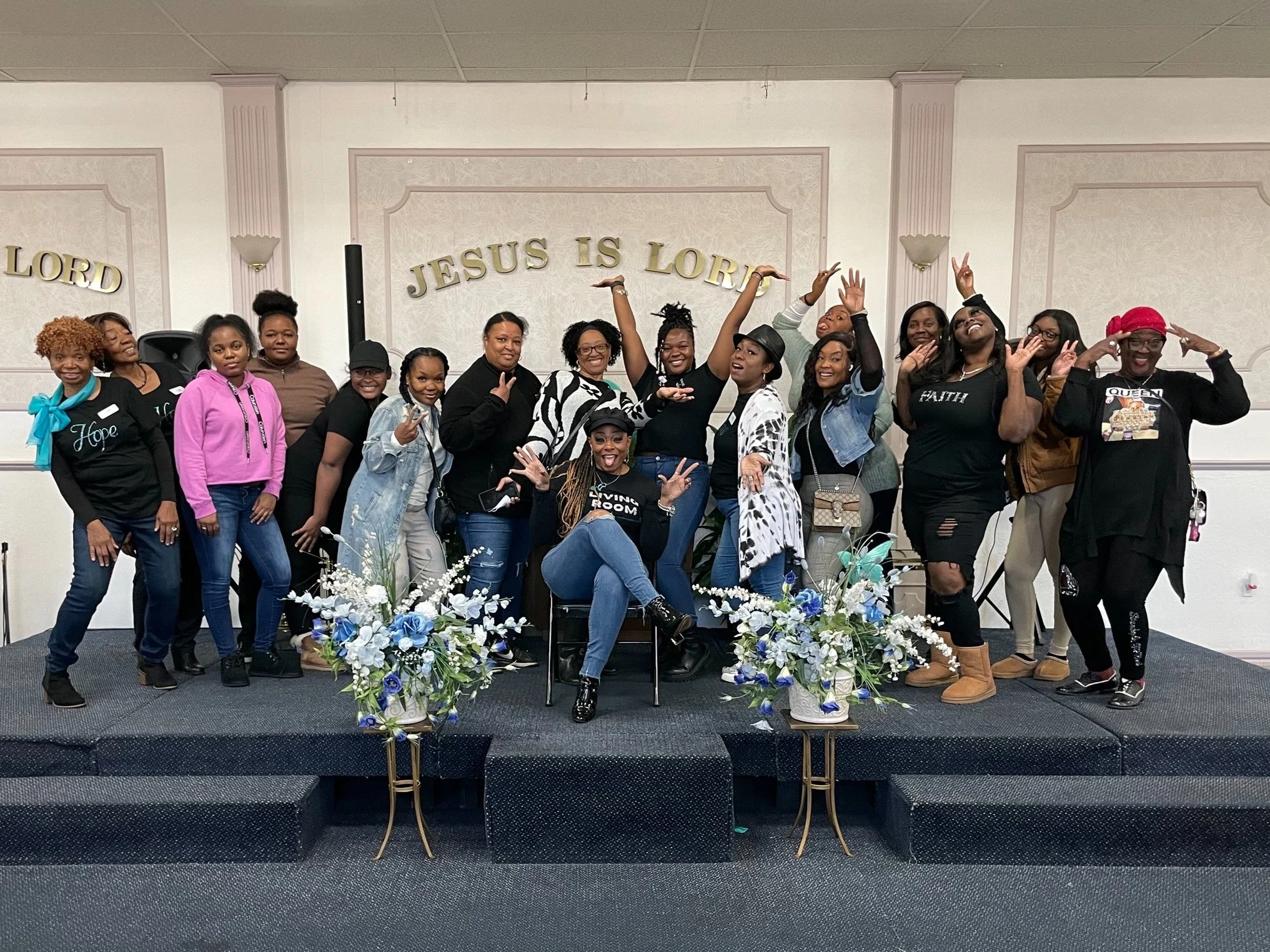 A group of women standing on a stage inside a church or community hall. The women are smiling, posing, and making peace signs or gestures of celebration. In the background, a wall with the words 'JESUS IS LORD' hung in gold letters is visible. The stage is decorated with a floral arrangement in front, featuring white, blue, and purple flowers. Some women are wearing black T-shirts with inspirational words, and others are in casual or semi-formal outfits.
