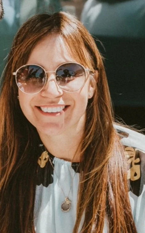 Smiling woman with long brown hair, bright red lipstick, wearing a white halter top with orange embroidery, outdoors with blurred trees in background.