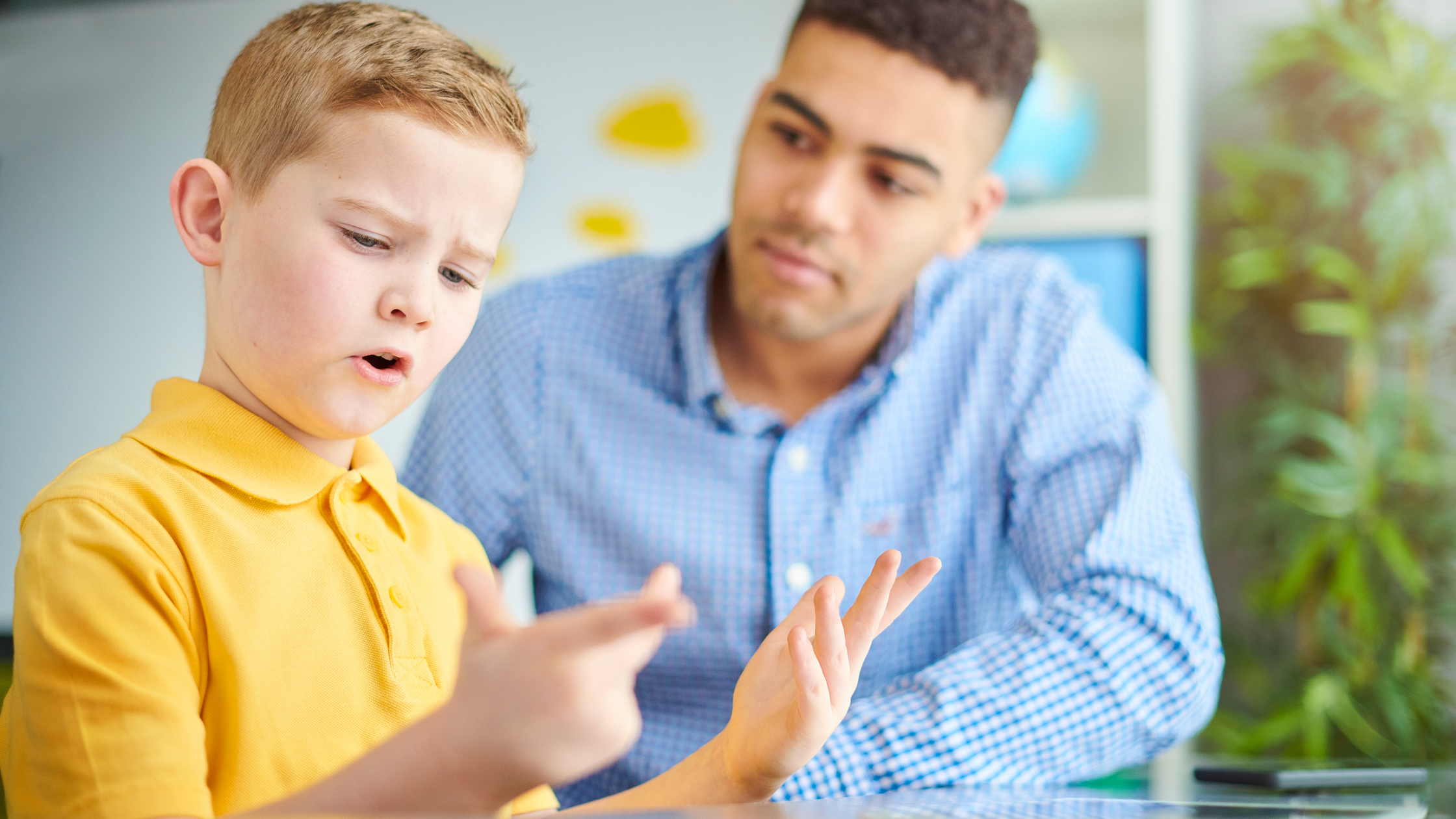 Boy in yellow t-shirt counting on his fingers with male teacher or parent in background watching.