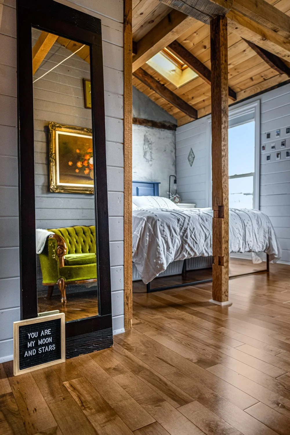 A cozy bedroom with a wooden ceiling, skylight, white shiplap walls, a large bed with white bedding, and a rustic exposed wood post. A tall mirror reflects a vintage green armchair and a framed floral painting. A small sign on the floor reads, "You a