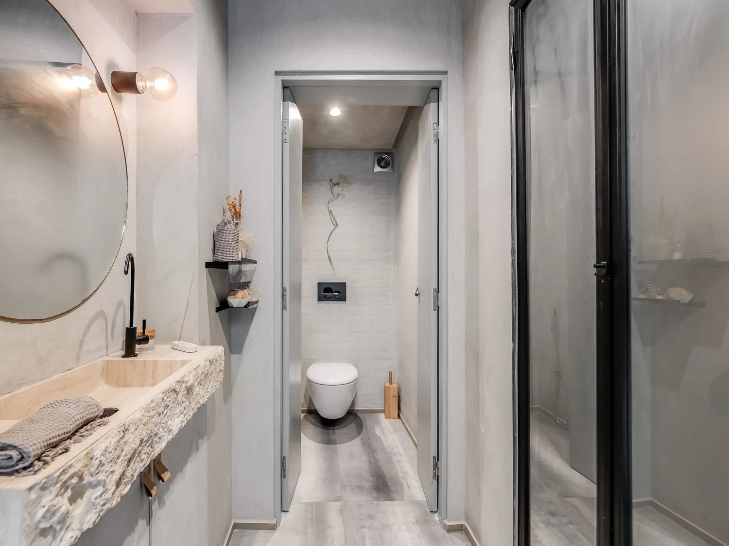 Modern bathroom with a wall-mounted toilet, a textured sink with a black faucet, a round mirror, a small black shelf with decor, a towel folded on the sink, and black-framed glass door cabinets on the right.