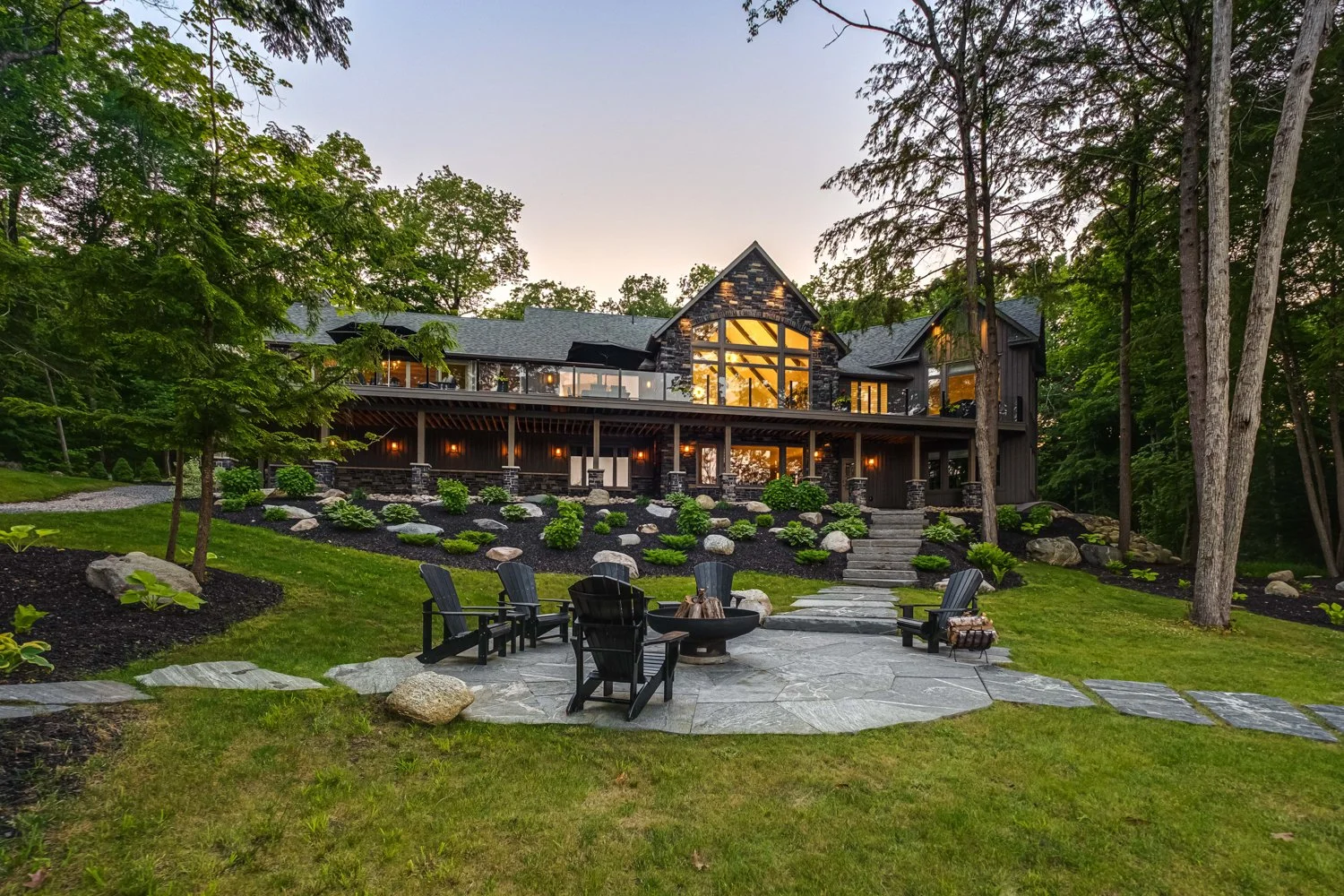 Large modern house with extensive windows surrounded by green trees, a landscaped garden with shrubs, rocks, and a stone patio with outdoor chairs and a fire pit.