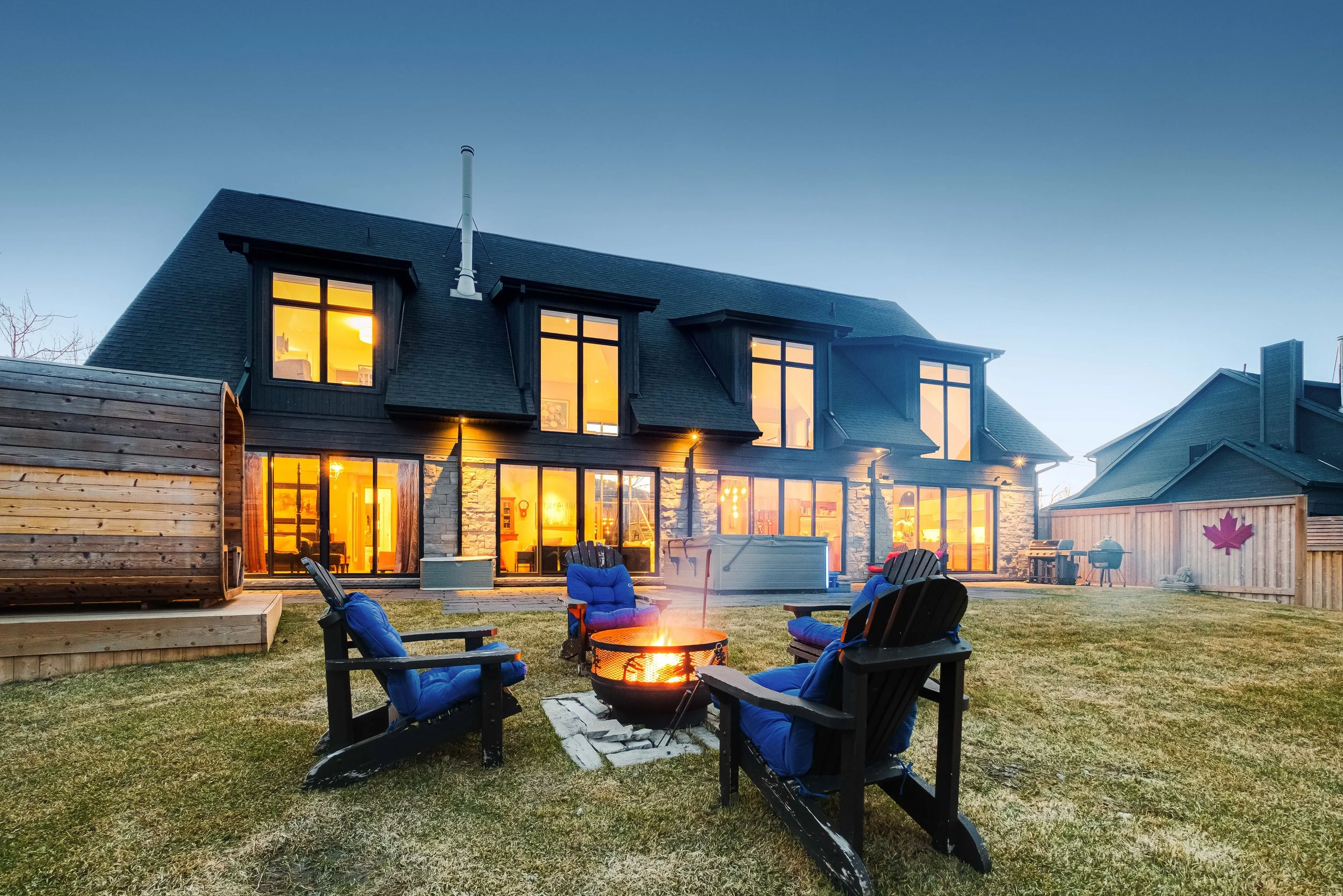 A backyard with a fire pit surrounded by four black chairs with blue cushions, a hot tub on the left, a large modern house with lit windows in the background, and a wooden fence with a pink maple leaf decoration on the right.