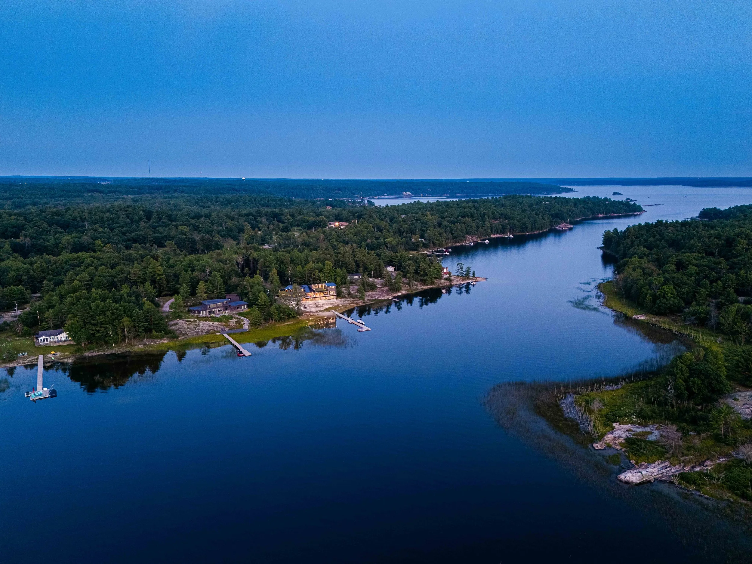 Aerial view of a river with several houses along the shoreline, surrounded by dense green trees, with docks extending into the water and a distant horizon under a blue sky.