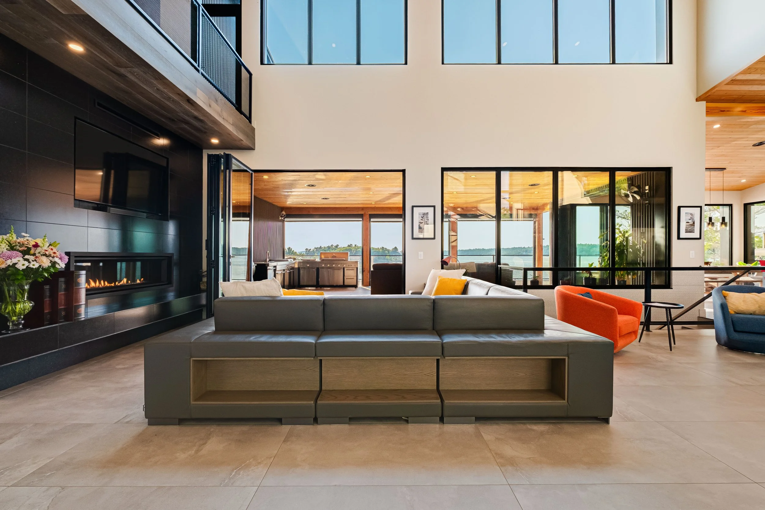 Spacious modern living room with large windows, colorful chairs, a fireplace, and a view of an outdoor deck and water.