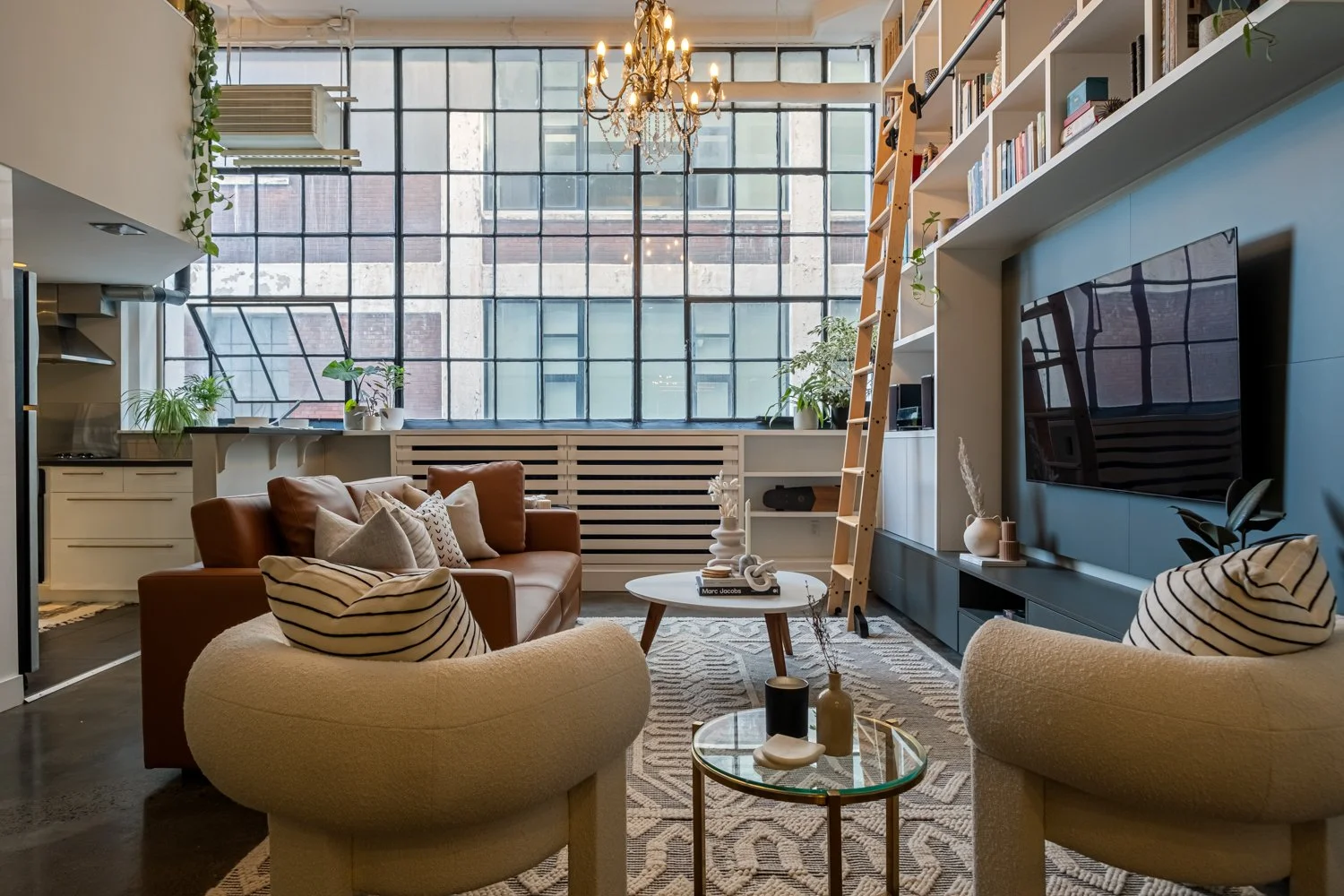 Modern living room with large window wall, brown leather sofa, two rounded beige armchairs with striped pillows, white coffee table, tall wooden ladder, bookshelves, plants, and a black TV mounted on a blue wall.