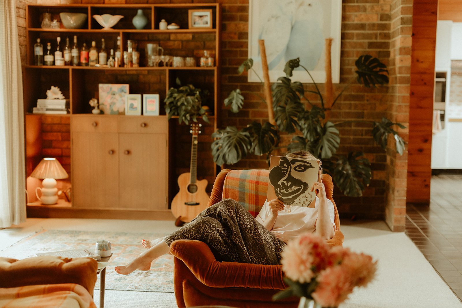 A woman sitting on an orange velvet sofa, in a 1960s house, covering her face with a book.