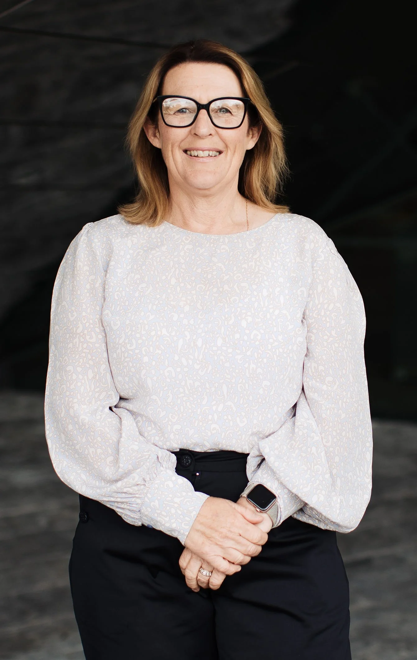 Sharon Williams, conference management team member at Composition. A woman with shoulder-length brown hair wearing large black glasses, a light-colored patterned blouse, black pants, and a smartwatch, smiling in front of a dark, blurred background.