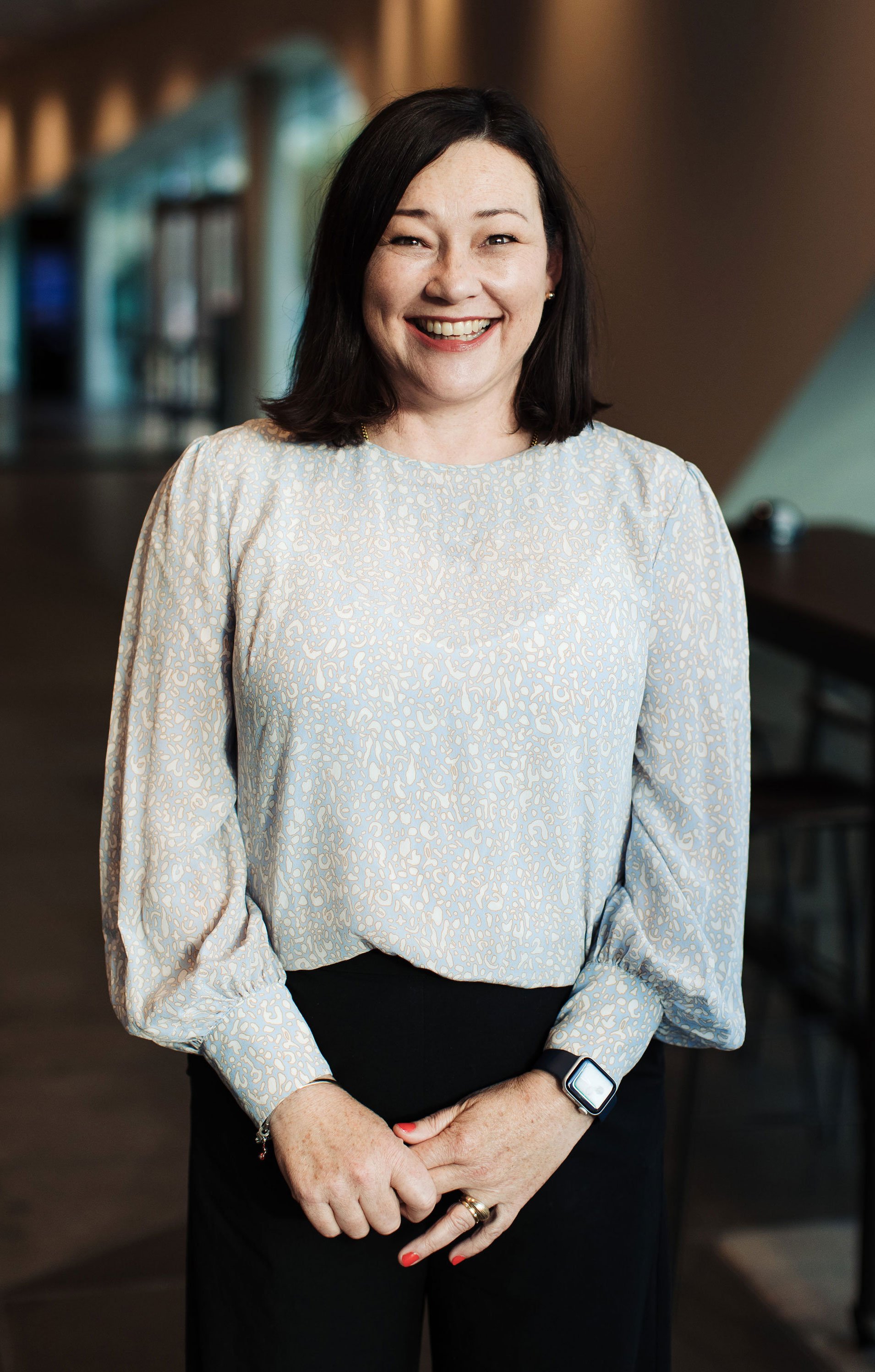Kelly Benison, conference management team member at Composition. A woman standing in an indoor setting, smiling, wearing a light-colored patterned blouse, black pants, and an smartwatch.