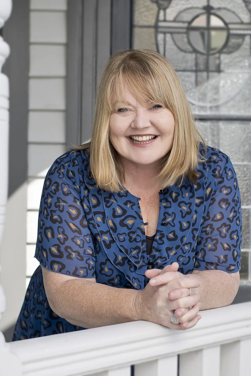 Nerida Ramsay, Director and Operations & Logistics Manager at Composition. Smiling woman with blonde hair wearing a blue leopard print blouse, leaning on a white railing with a house and stained glass window in the background.