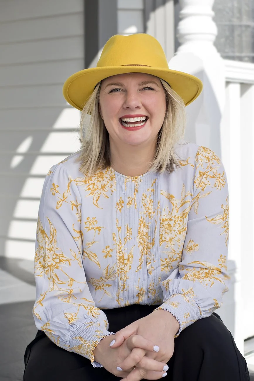 Arna Wahl Davies, Director and Client & Communications Manager at Composition. A woman with blonde hair, smiling broadly, wearing a yellow hat and a white blouse with yellow floral patterns, sitting outdoors near a porch with white railings.