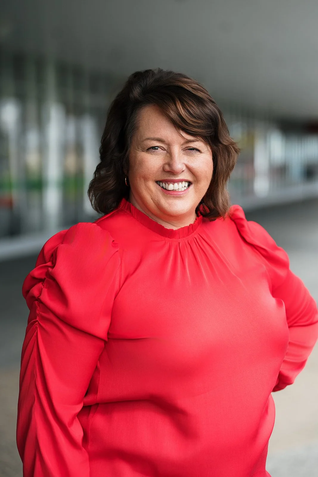 Jen Henshaw, conference and event management team member at Composition. A woman with shoulder-length brown hair smiling in a red blouse outdoors with a modern building in the background.