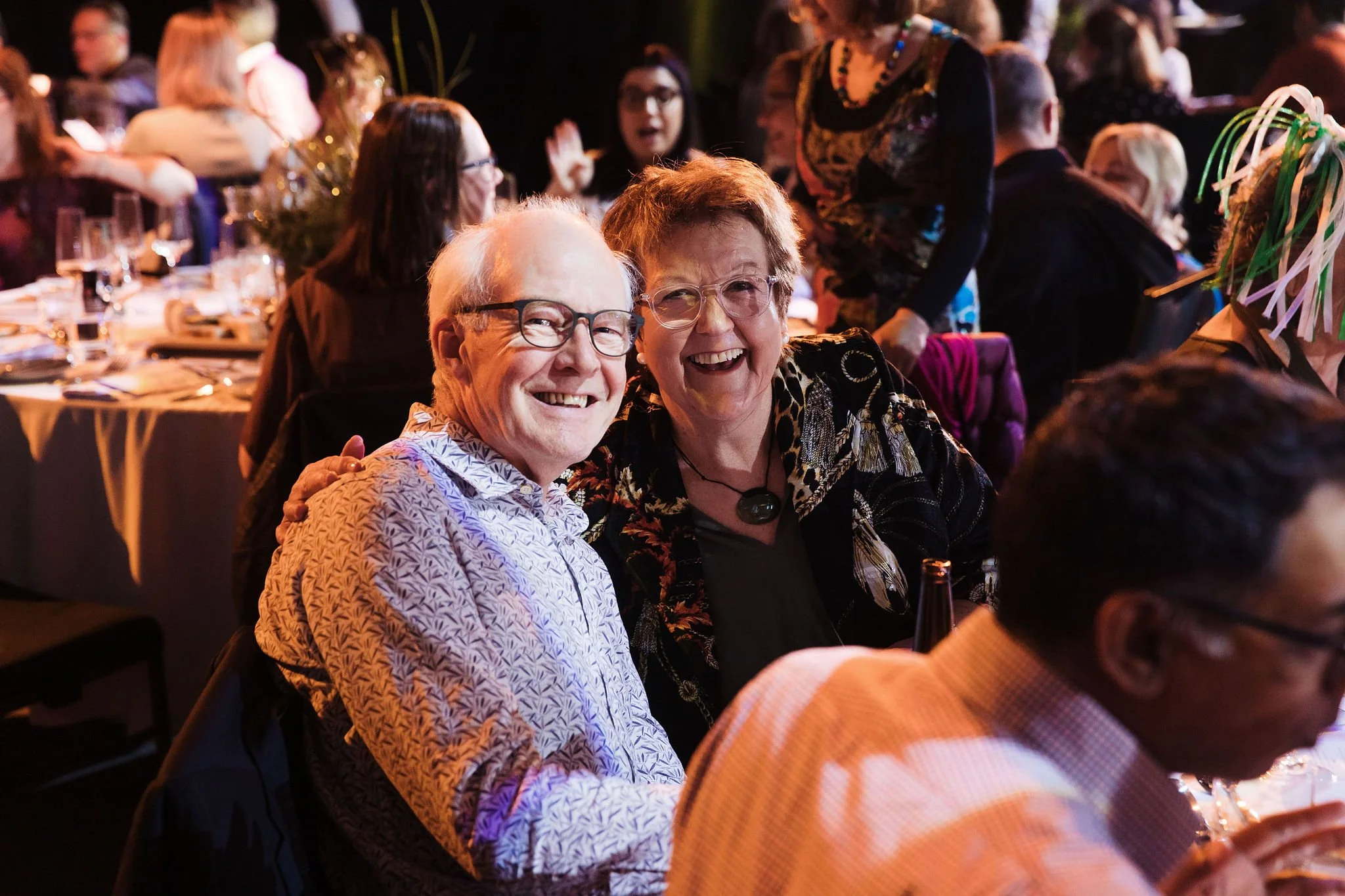 Delegates at the Hospice New Zealand conference, one of the conferences managed by Composition. Two smiling older adults, a man and a woman, sitting together at a crowded event, with a lively background of other guests and tables