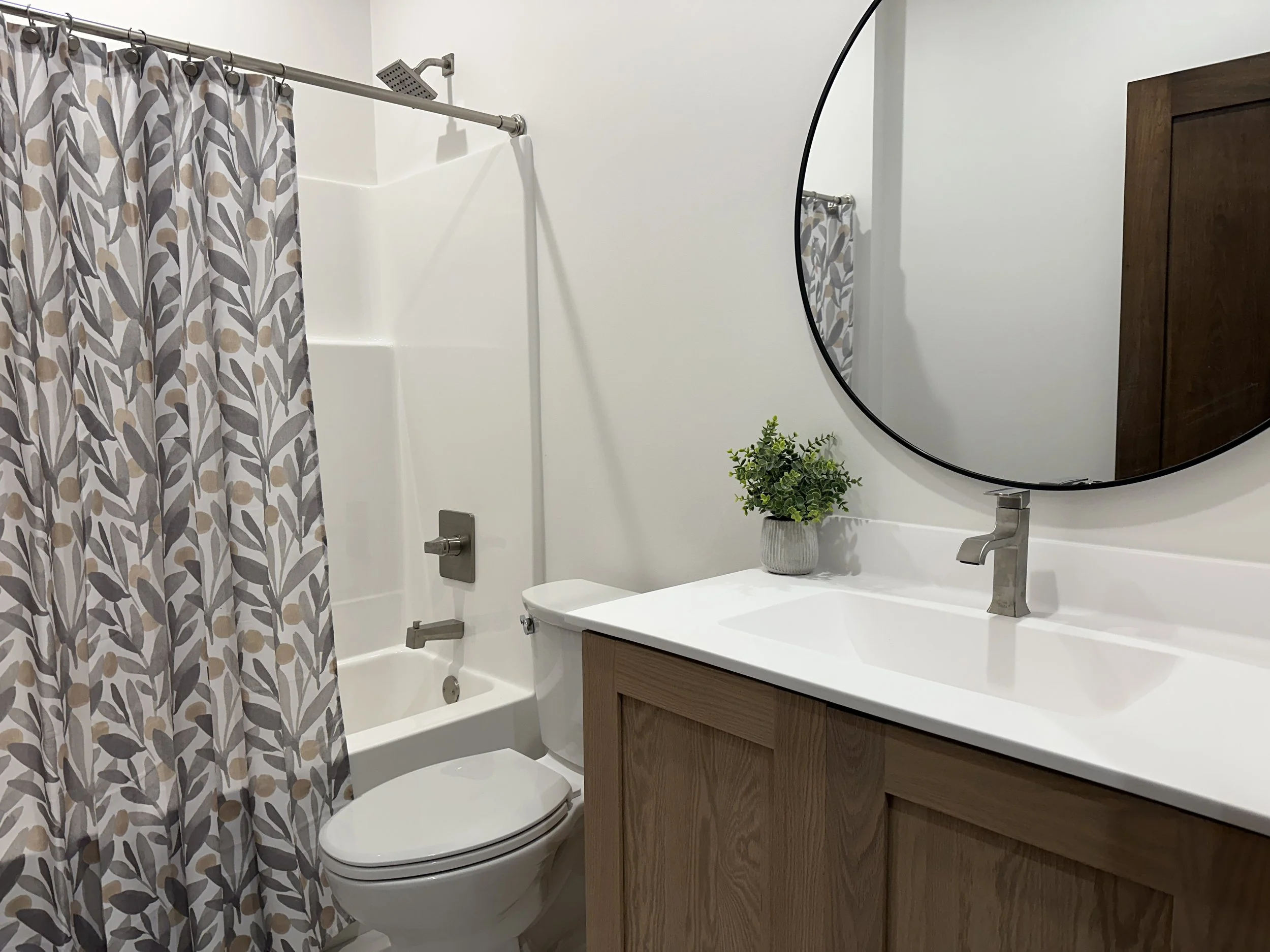 Modern bathroom with white walls, a shower with a patterned curtain, a toilet, a wooden vanity with a white countertop, a gray vase with green plants, and a large round mirror.