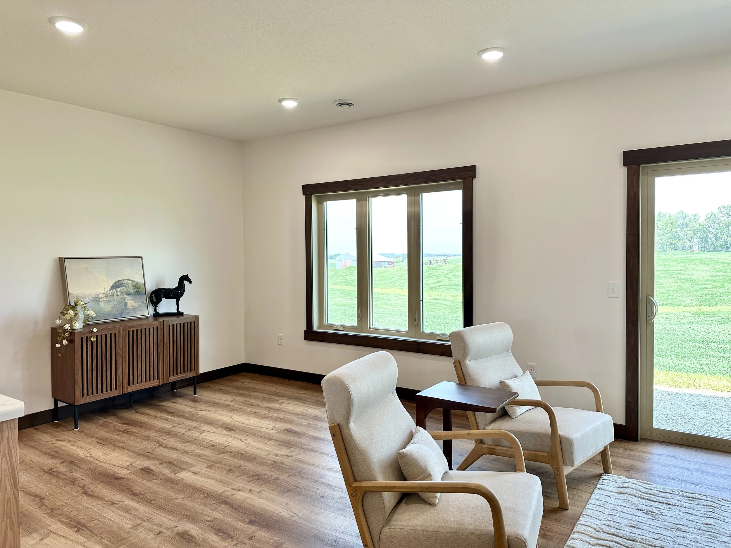 Spacious living room with two cream-colored armchairs, a small wooden side table, large windows, a glass sliding door showing a green field outside, a wooden sideboard with artwork, a horse sculpture, and decorative flowers, with a light wooden floor