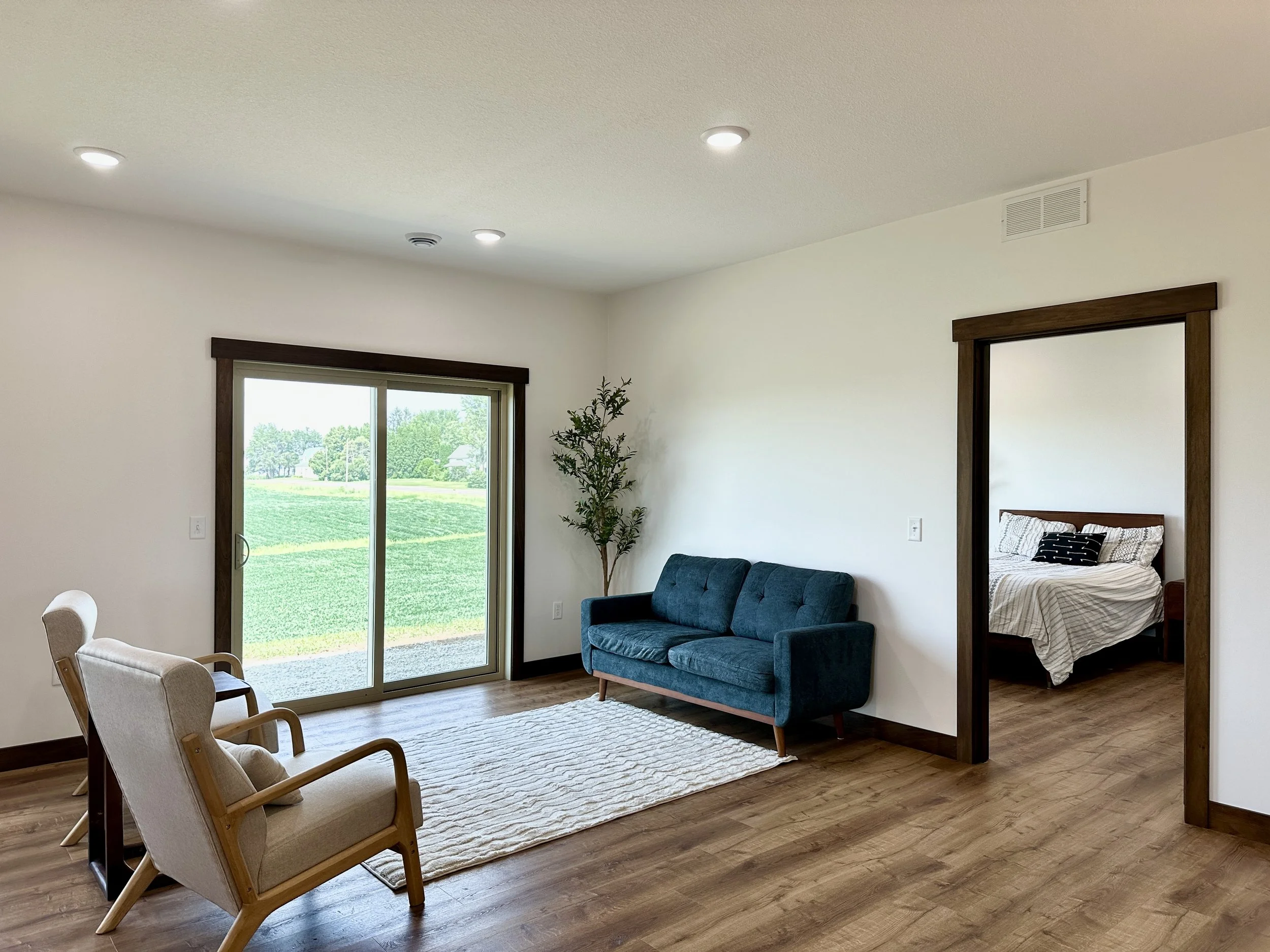 A living room with a sliding glass door leading outside, a blue sofa, two beige armchairs, a white rug, a potted plant, and a view of a green lawn outside. A doorway leads to a bedroom with a bed and striped bedding.