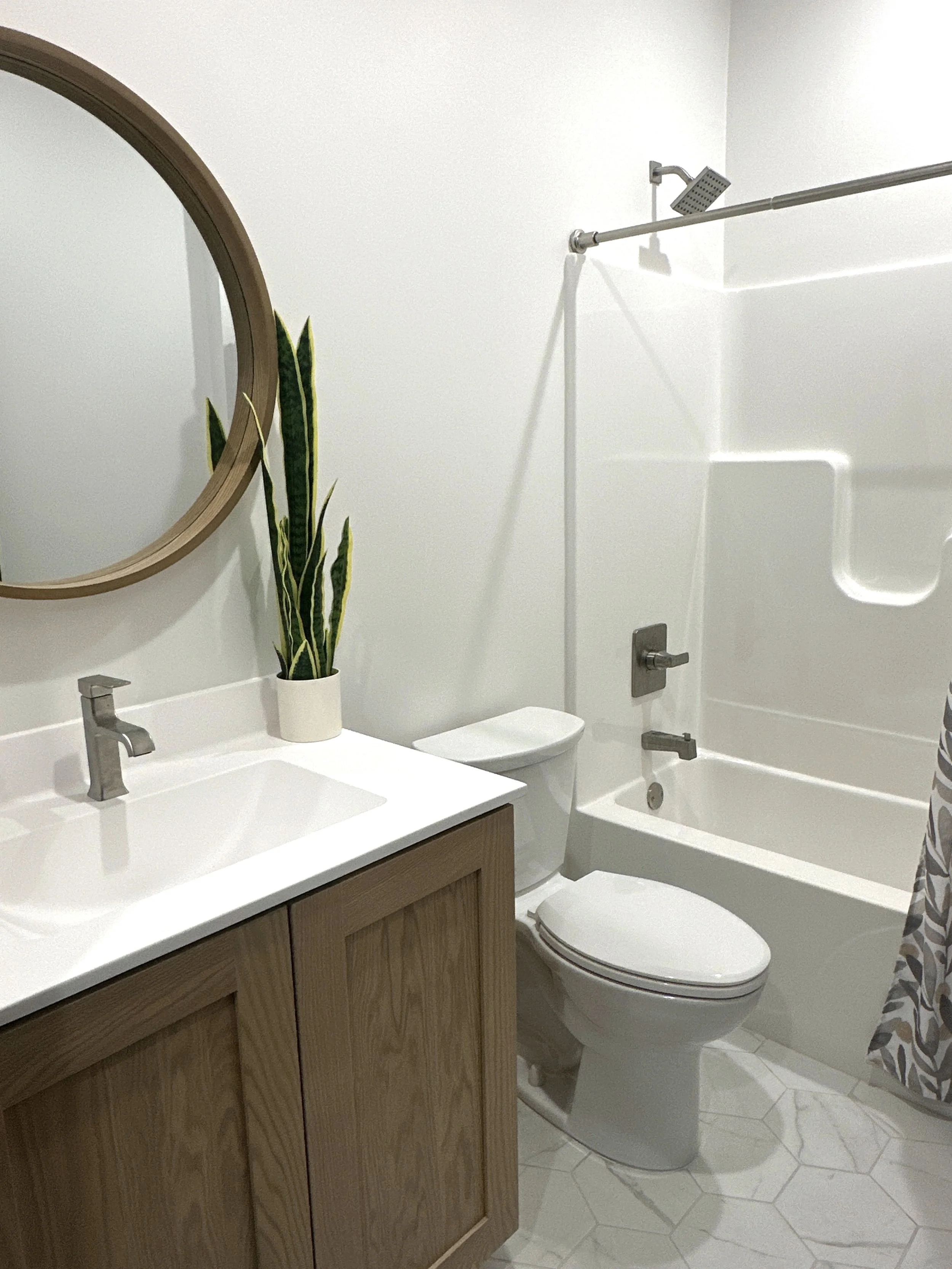 Modern bathroom with a wooden vanity, white sink, round mirror, potted snake plant, toilet, bathtub with showerhead and curtain.