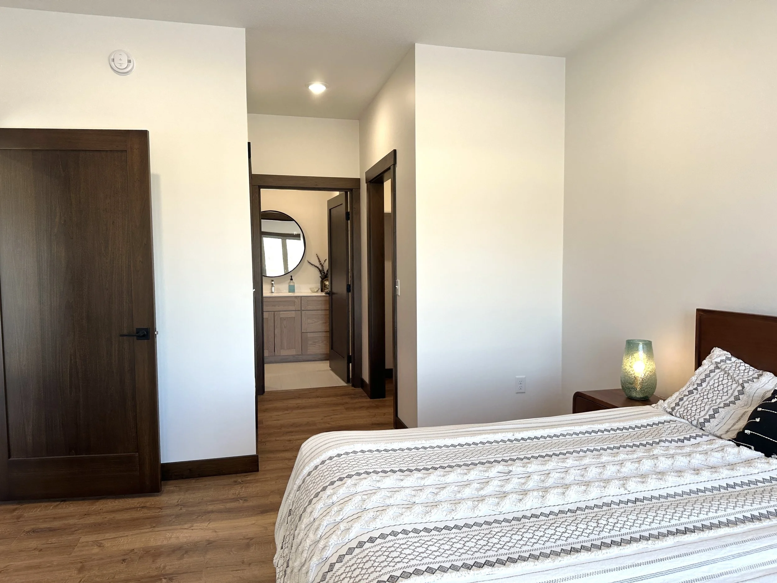 A bedroom with a bed, nightstand, and lamp, with an open door leading to a bathroom with a vanity and mirror.