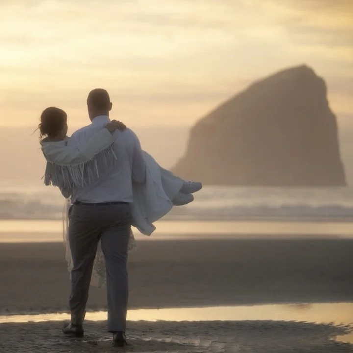 A groom carrying his bride along the beach toward a sea stack at sunset