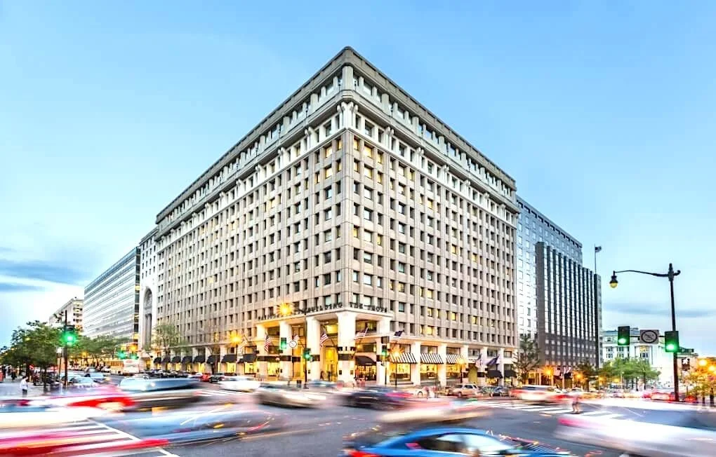 City street scene with a large multi-story building, cars moving, traffic lights, and pedestrians during dusk