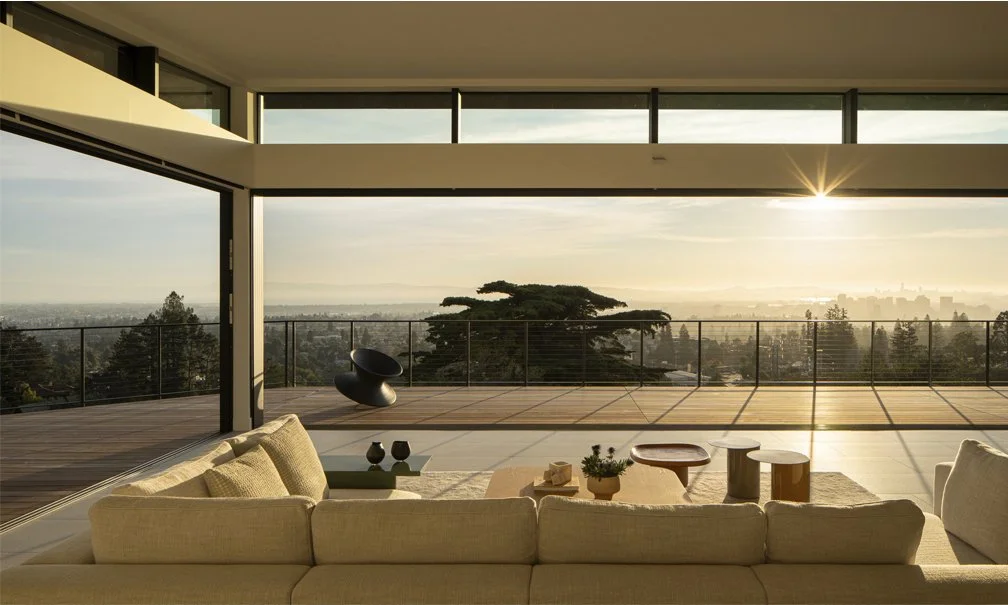 Light-filled living room with floor-to-ceiling sliding glass walls open to a terrace, cream sectional sofa, and panoramic views of the Bay at sunset.