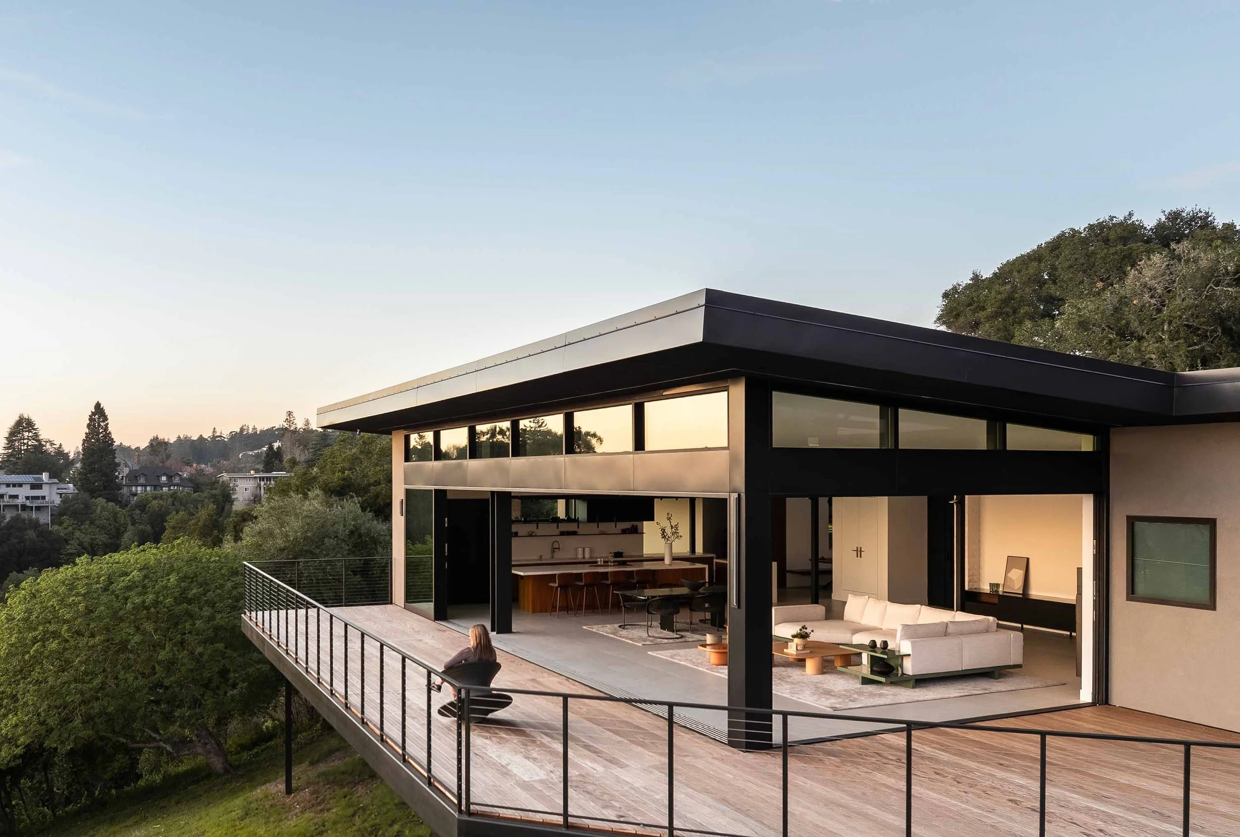Aerial dusk view of the Oakland Hills home with walls fully open to the terrace, revealing the kitchen, dining, and living areas floating above a native oak landscape.