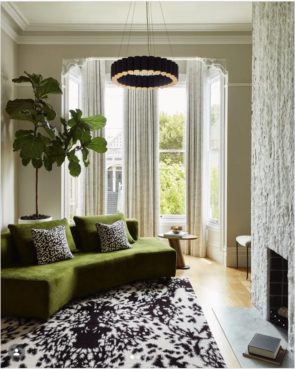 Victorian-era living room in Pacific Heights featuring a sculptural green velvet sofa, bold patterned rug, textured stone fireplace, bay windows with linen drapes, and a dramatic black chandelier.