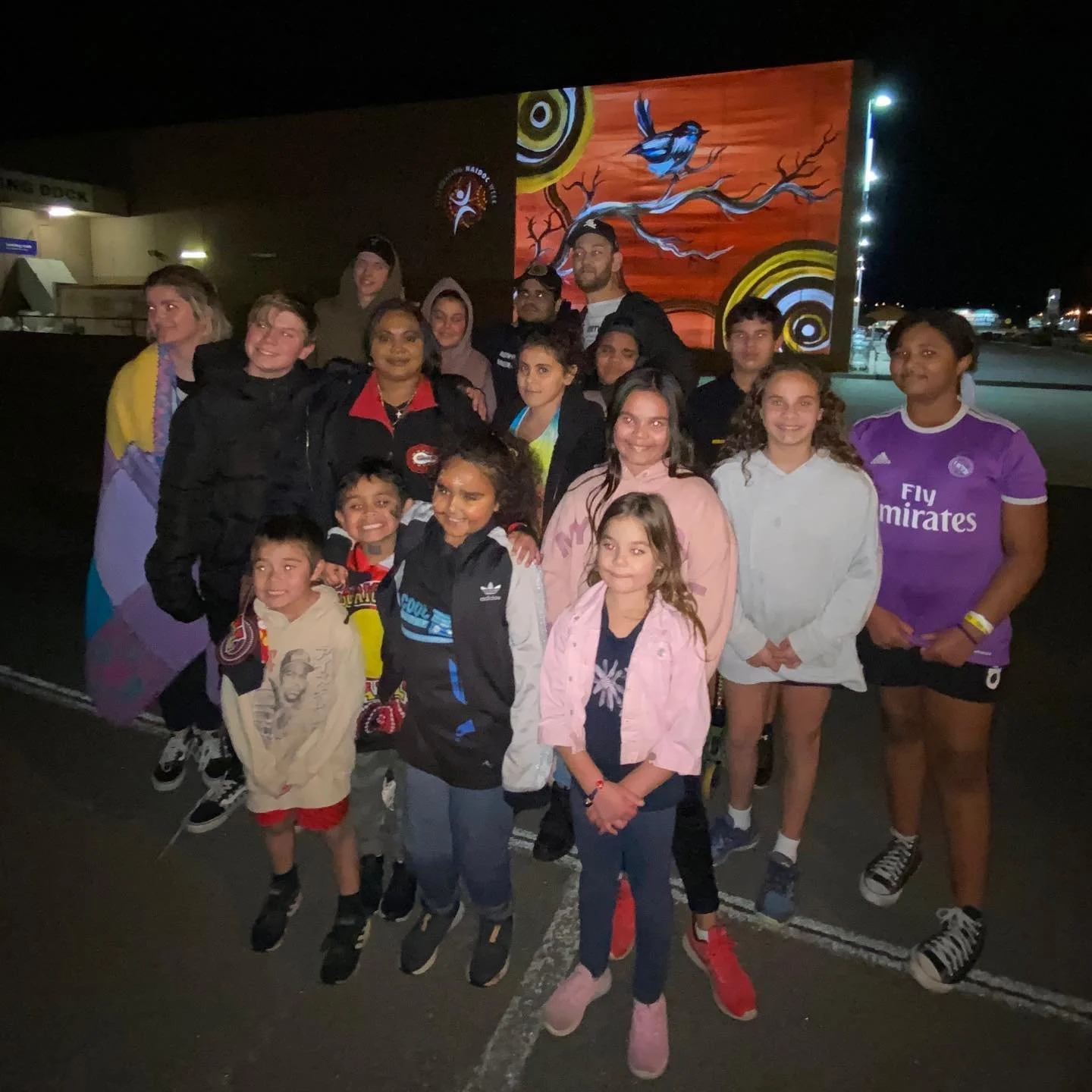 Group of children and adults gathered outdoors at night near a large, colorful mural with nature and bird designs.