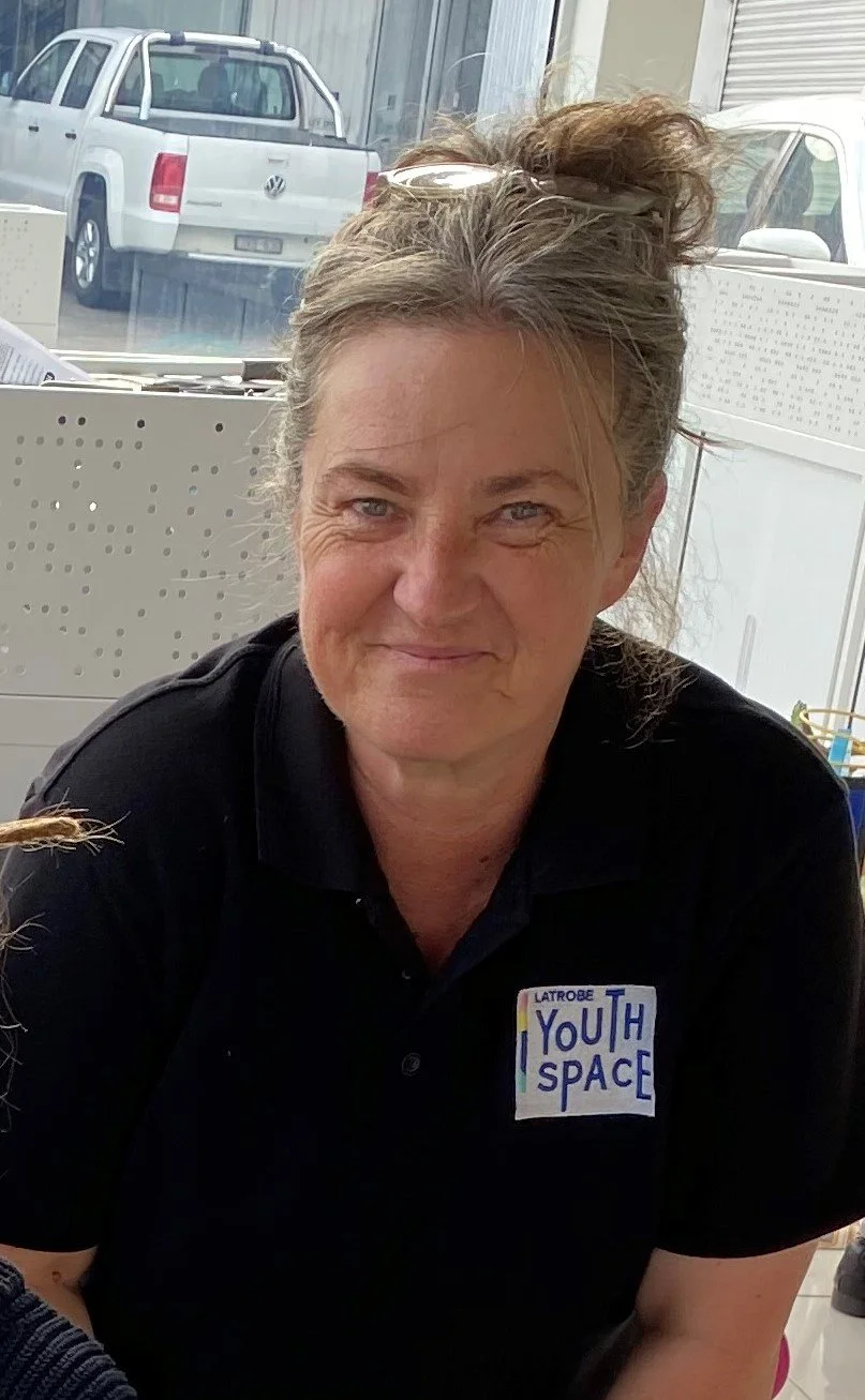 A woman with light brown hair in a messy bun, wearing a black polo shirt with a white name tag that says 'YOUTH SPACE.' She is sitting indoors near a window with parked trucks visible outside.