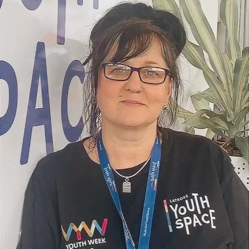Woman with dark hair, glasses, and a necklace, wearing a black t-shirt and a lanyard, sitting in front of a plant and a banner that says 'Youth Space'.