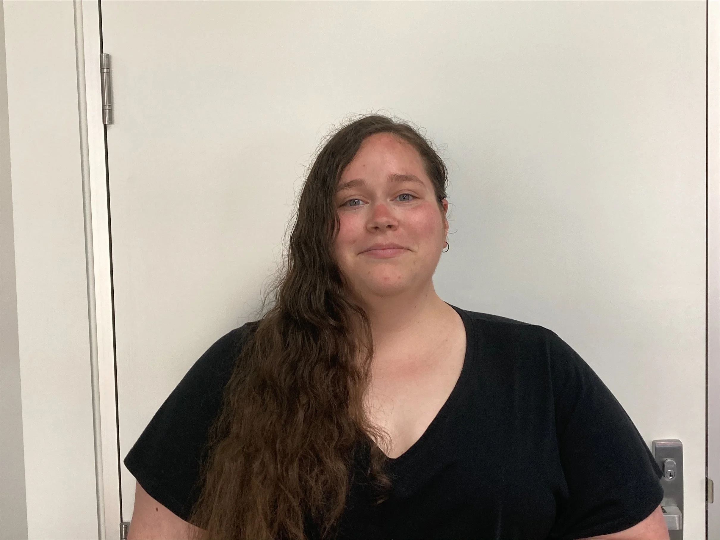 A young woman with long, wavy brown hair and fair skin smiling, standing in front of a plain white wall and a door.