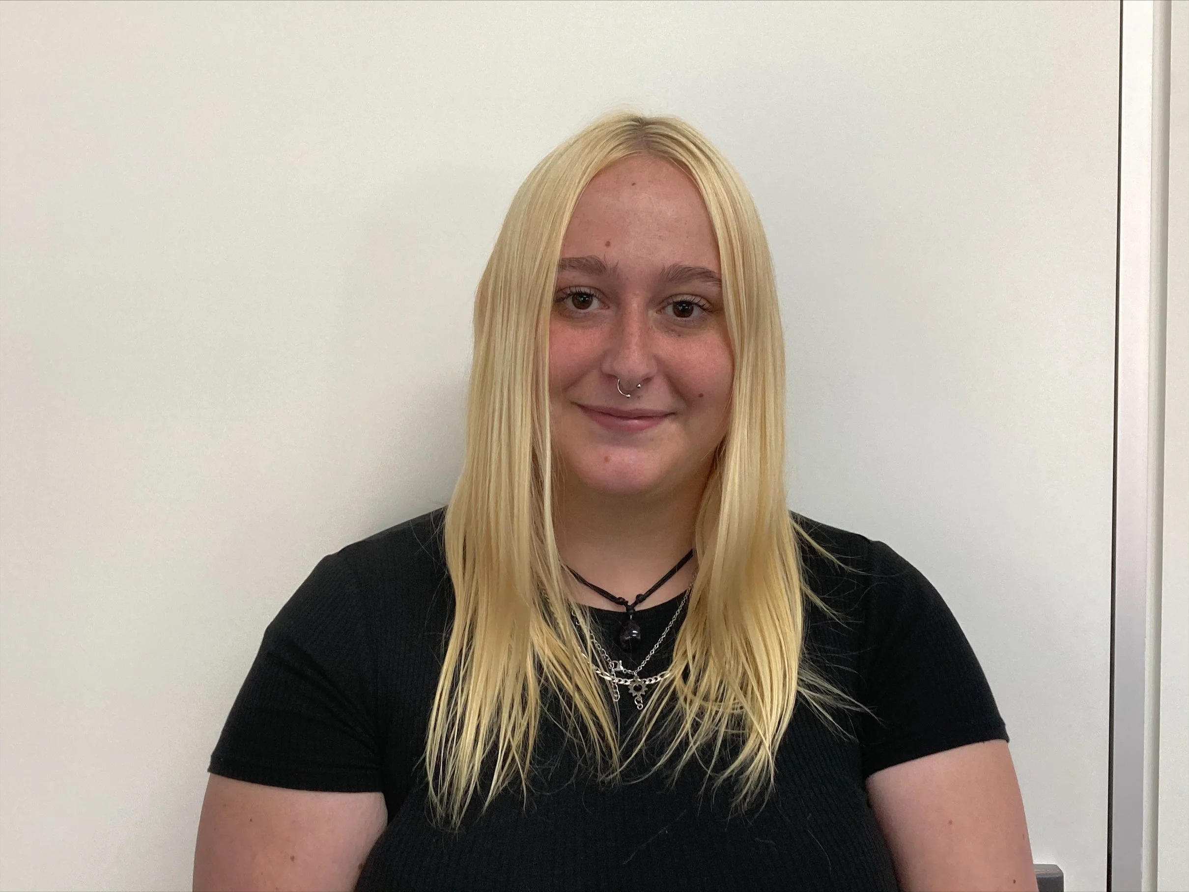 A young woman with long blonde hair, wearing a black shirt and layered necklaces, smiling at the camera against a plain white wall background.