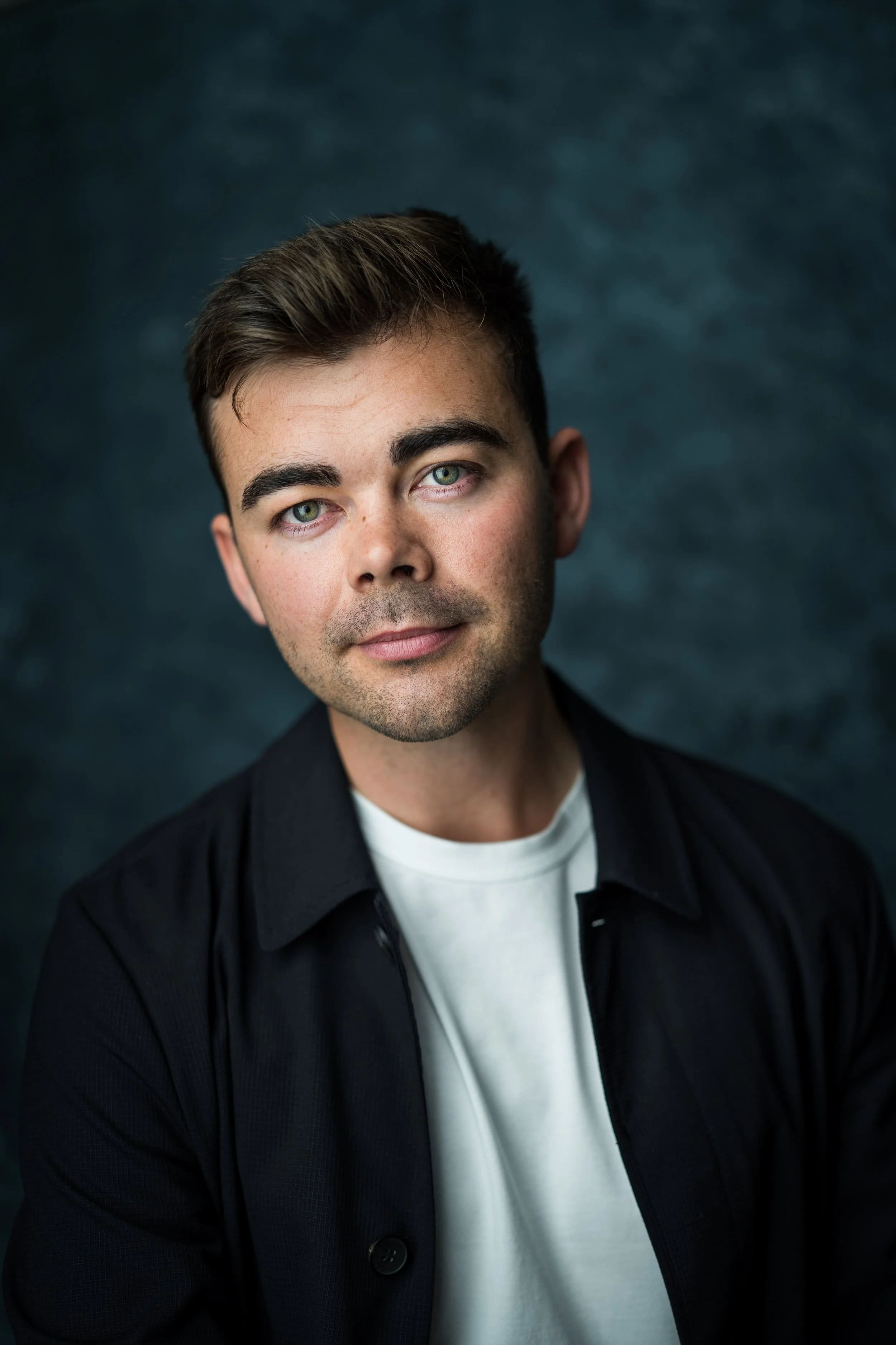 Studio headshot of Andrew McNicol wearing a dark jacket and white top against a blue textured background.