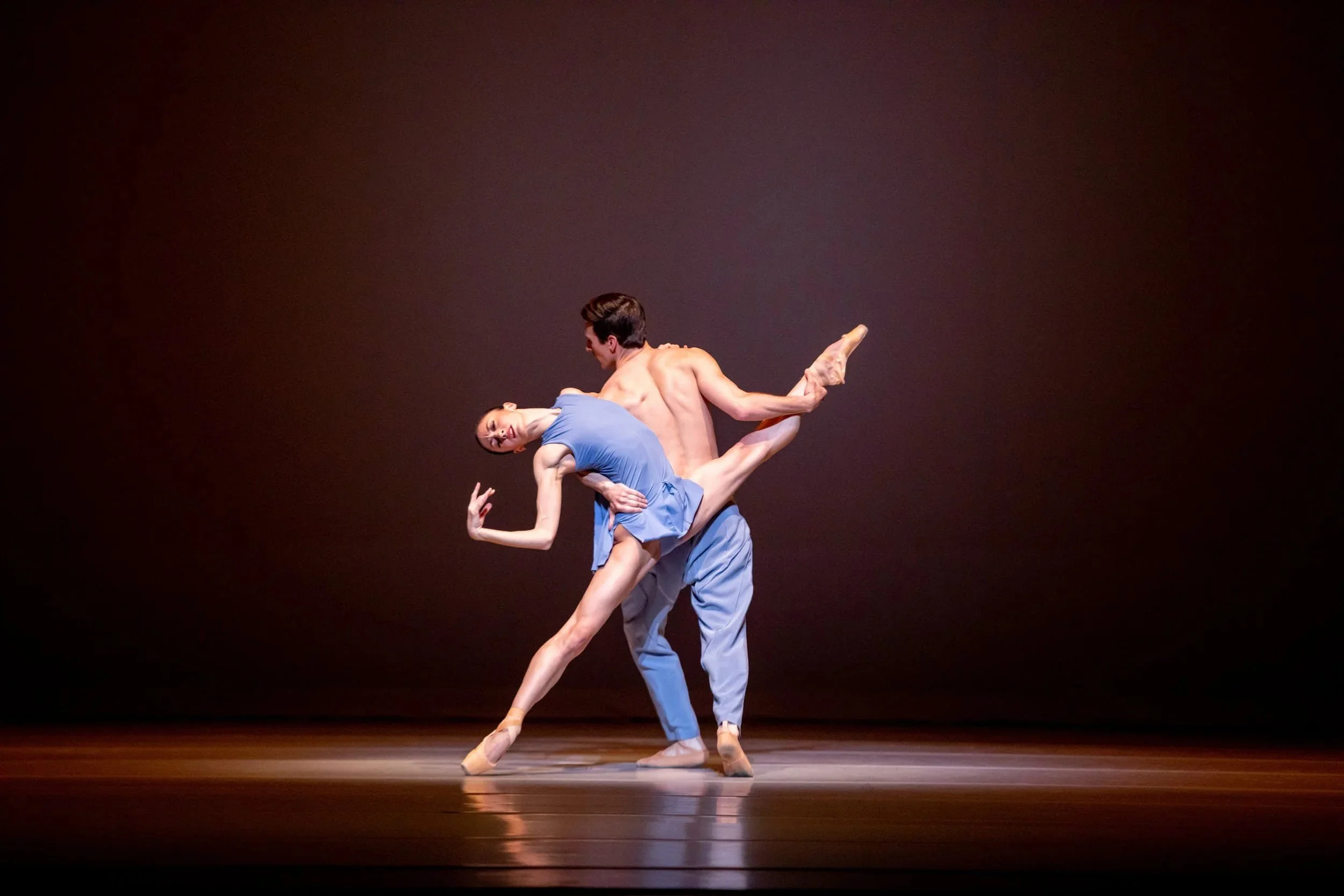 Two ballet dancers perform a lift on stage; a dancer in a blue dress leans back as her partner supports her under warm light.