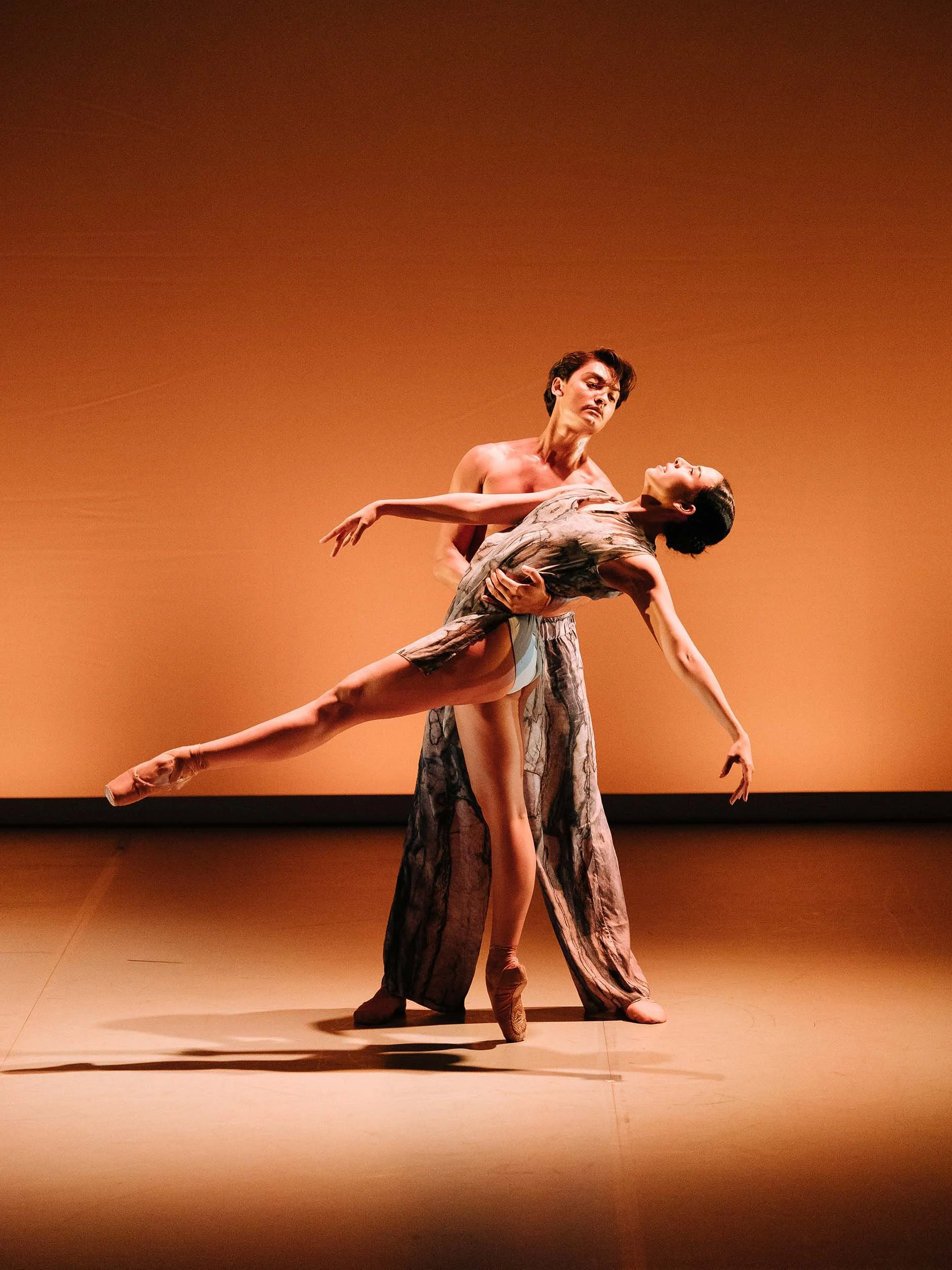 Ballet duet: a dancer is supported in a deep backbend with one leg extended, against a warm orange backdrop.