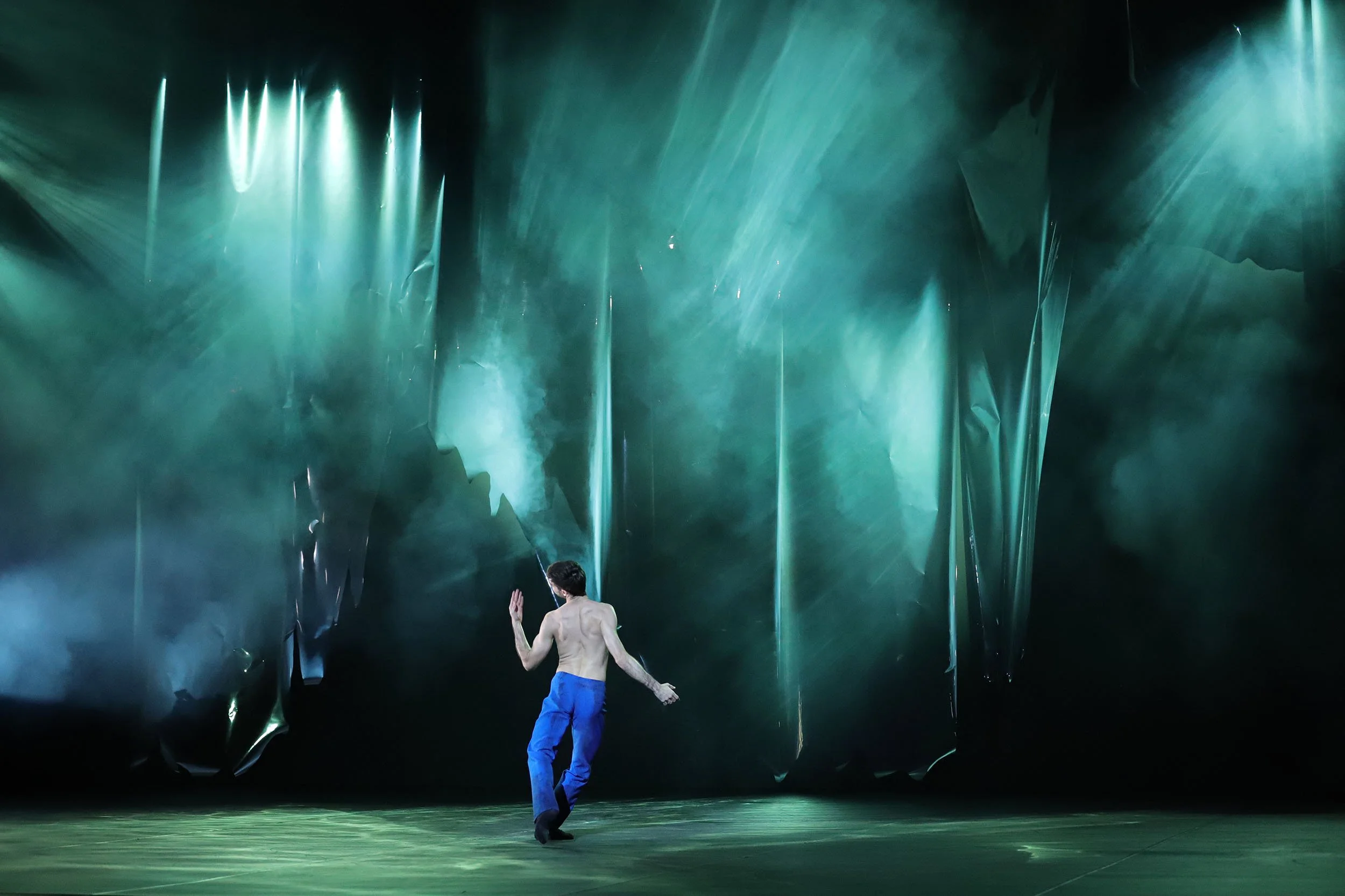 Solo dancer in blue trousers moves across a stage beneath dramatic green-blue lighting and hanging reflective set pieces.