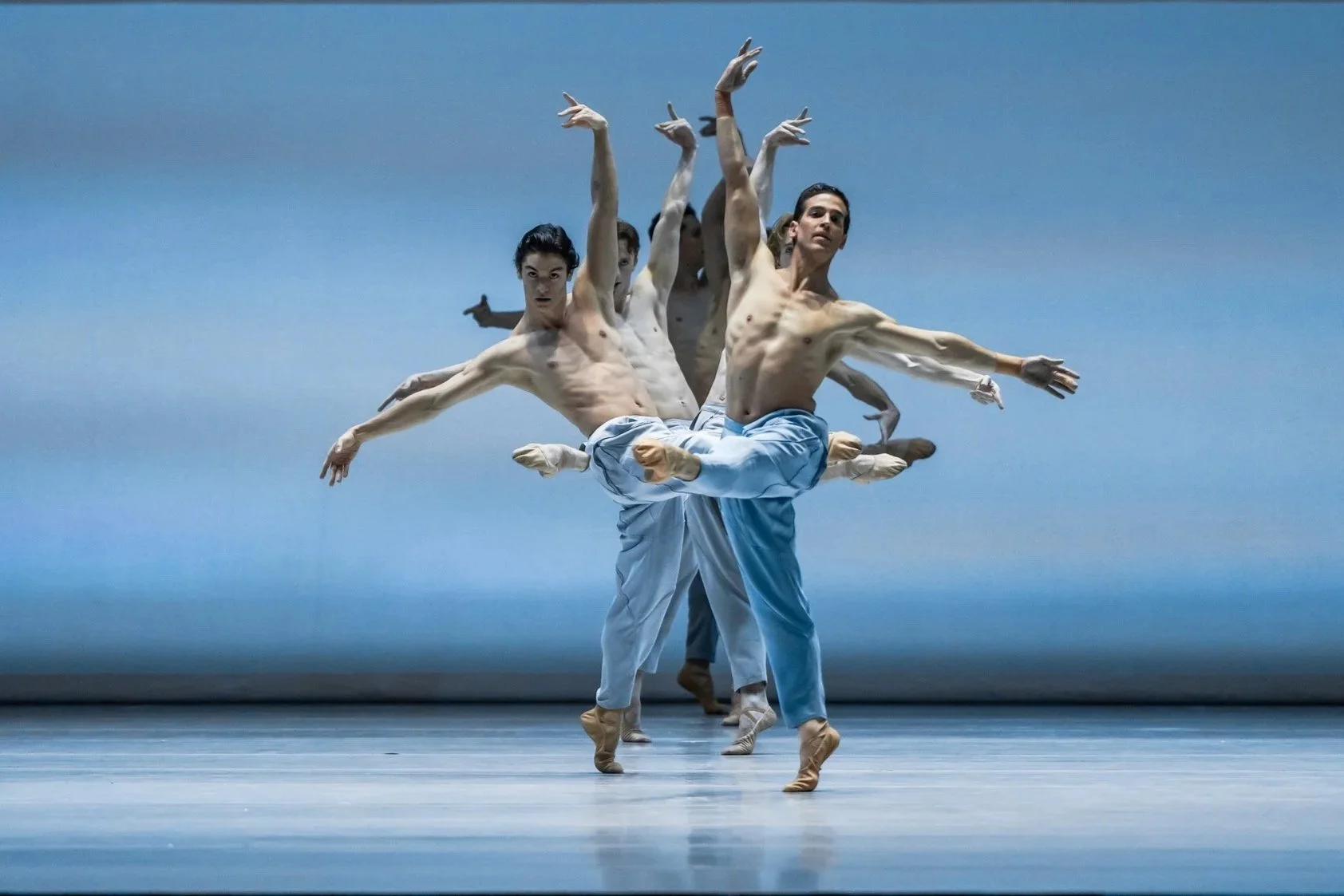 Male ballet dancers in blue trousers perform in formation on stage, arms extended against a pale blue backdrop.