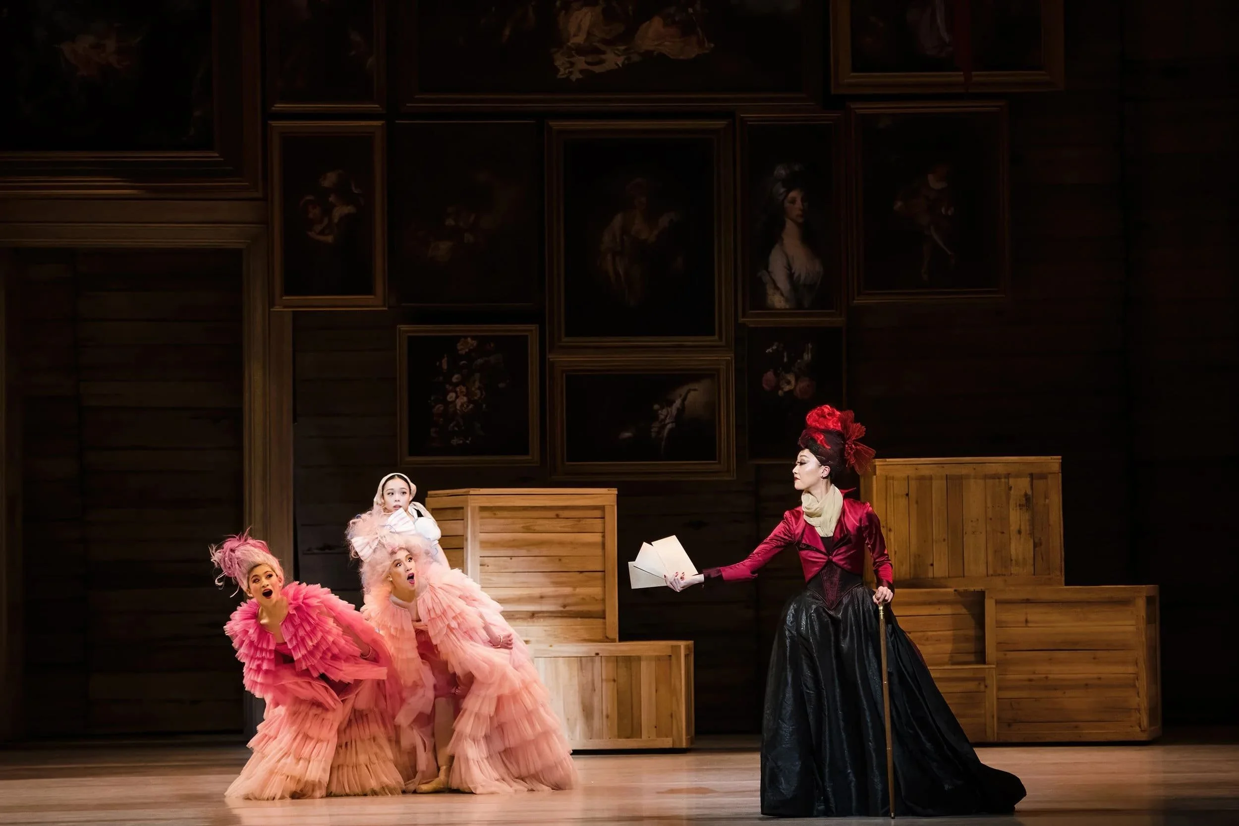Stage scene with two performers in pink costumes and a child, watched by a performer in a red top and black skirt holding a letter.