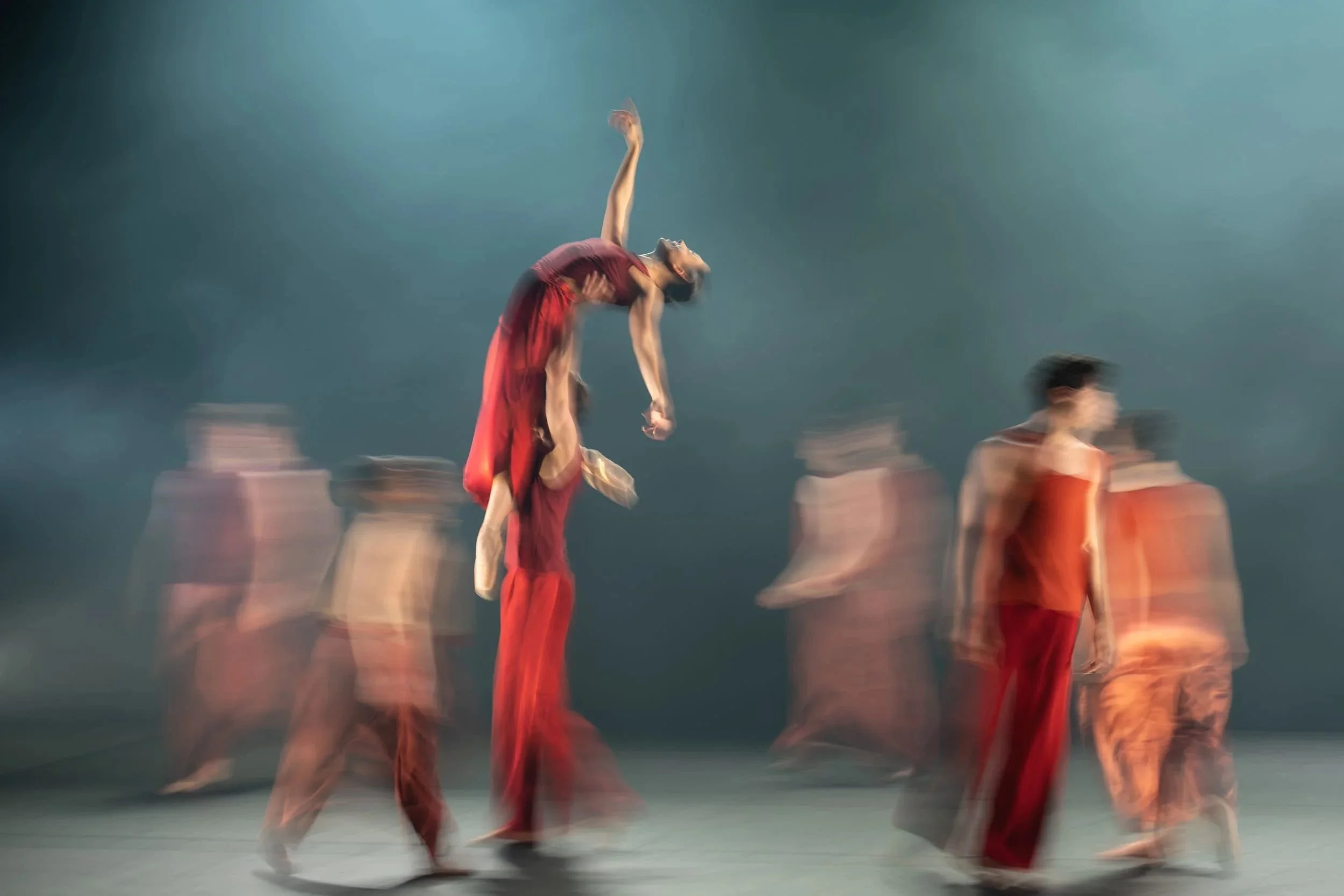 Contemporary dance ensemble in motion; a dancer in red is lifted and arched back under hazy blue stage light.