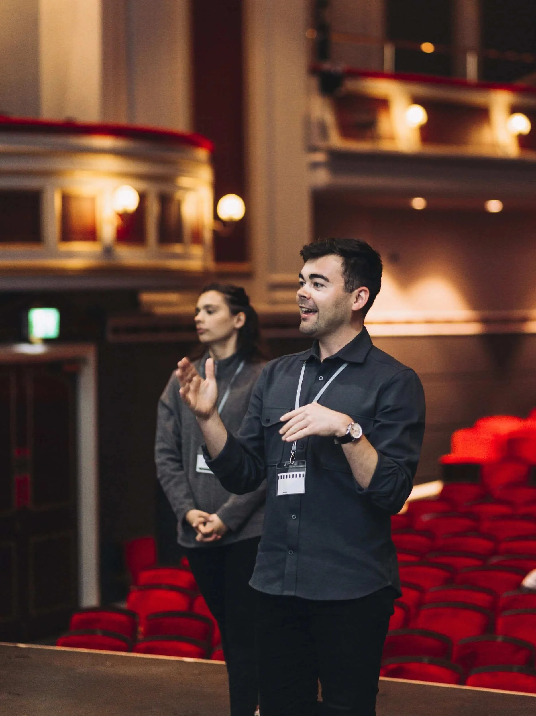 Andrew McNicol speaks in a theatre auditorium, gesturing as he addresses a group with red seats behind him.