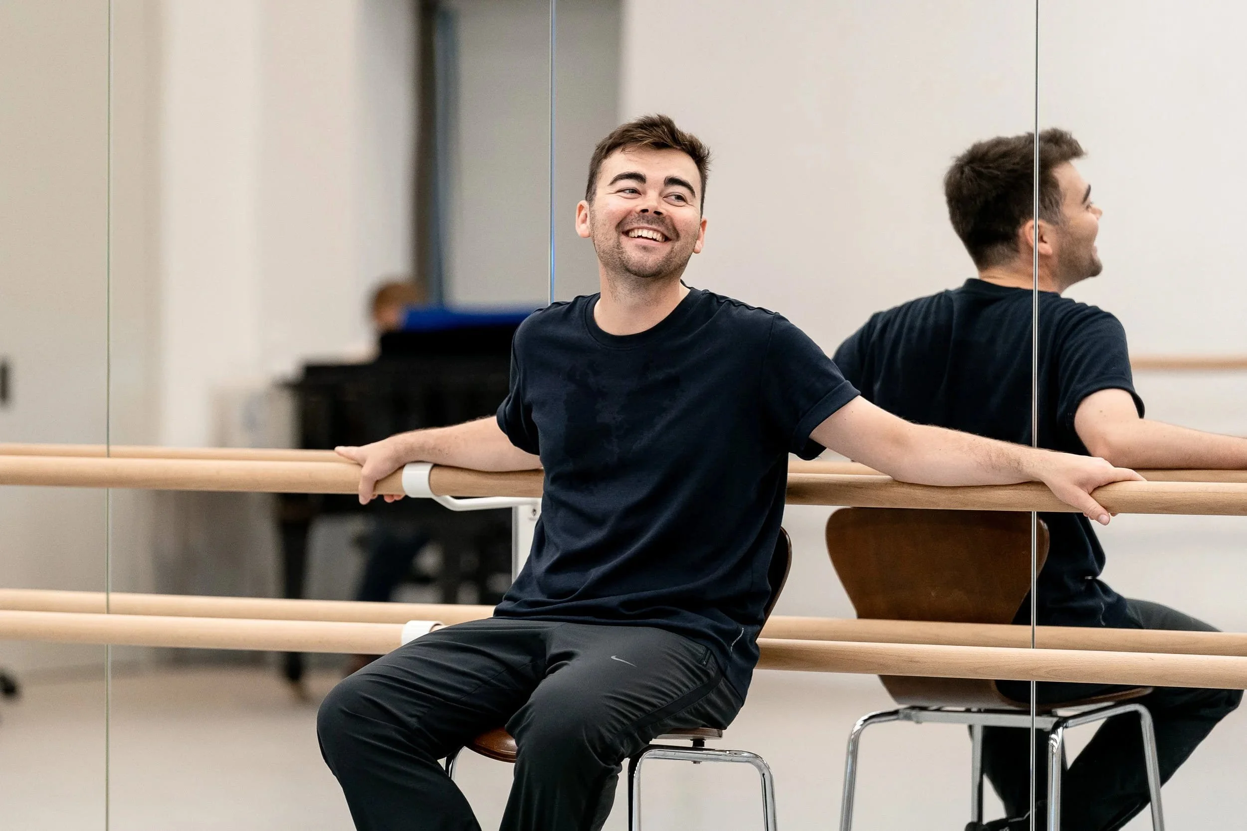 Andrew McNicol smiles while sitting by a ballet barre in a dance studio, reflected in the mirror behind him.
